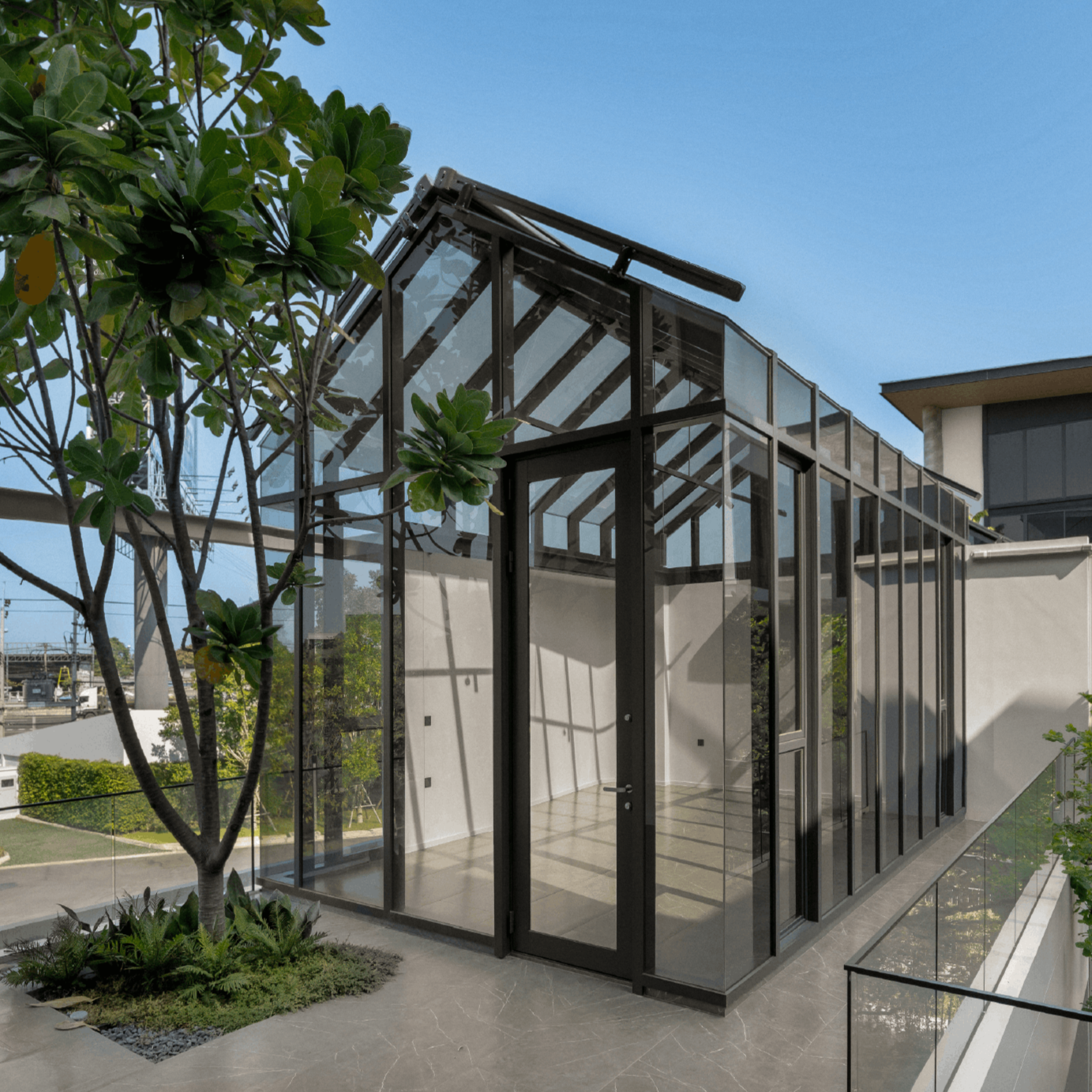 Modern glass greenhouse with black framing, visible interior shelves, and a door, next to a garden with a small tree and green plants under a clear blue sky.