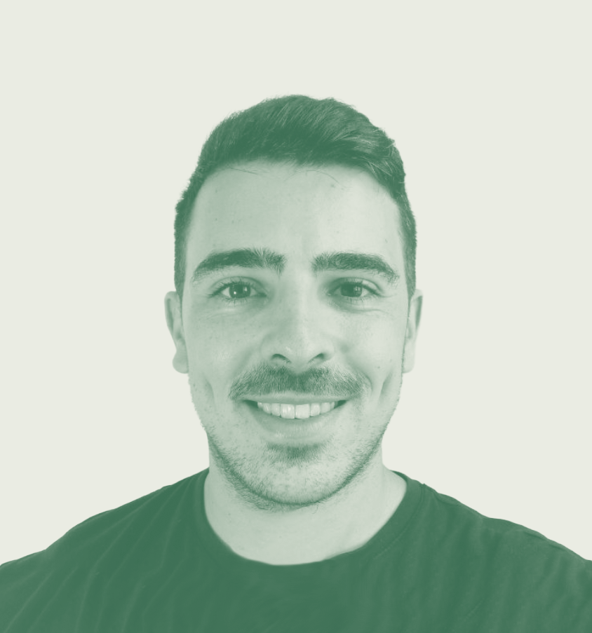 Close-up of a young man with dark hair, thick eyebrows, a mustache, smiling, wearing a black shirt against a plain background.