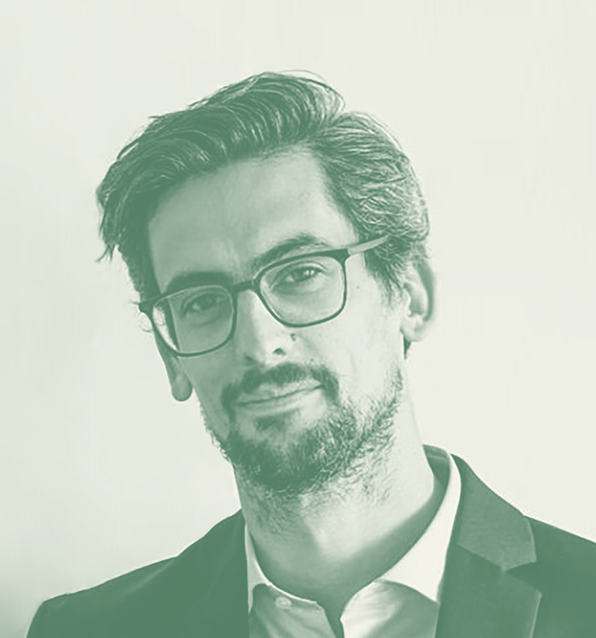 A portrait of a man with glasses, dark hair, and facial hair, wearing a suit jacket and white shirt, against a light background.