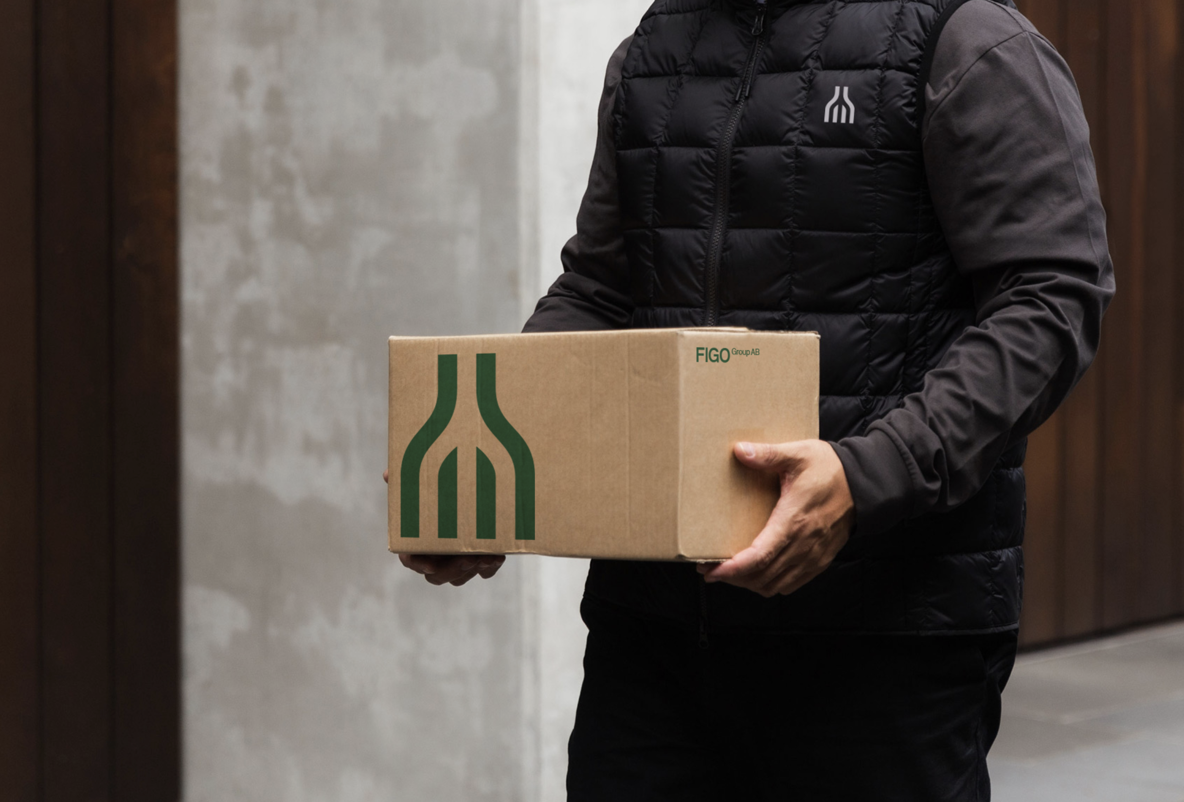 Person holding a cardboard box with a green logo, wearing a black puffer jacket, standing outdoors.