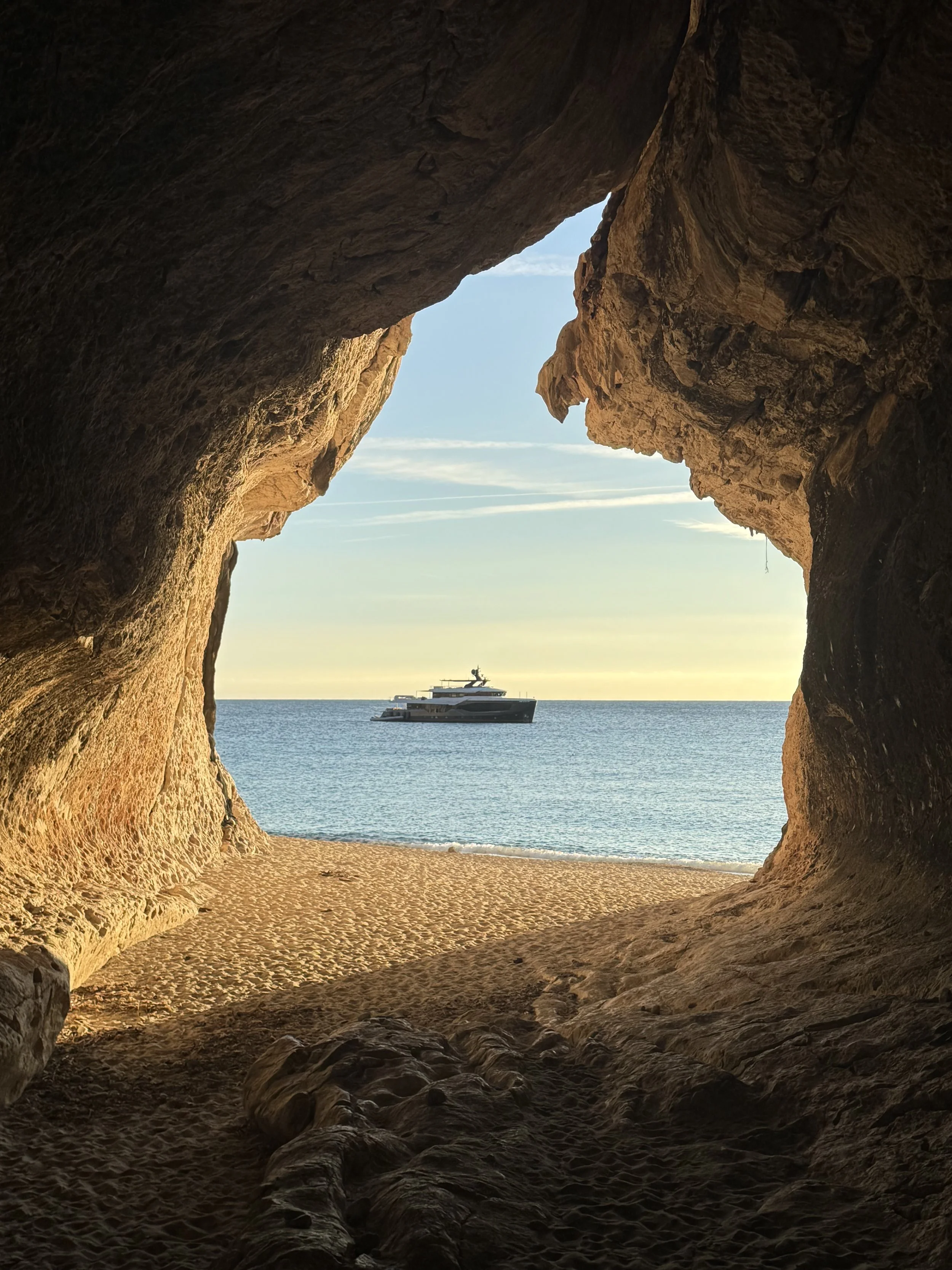 View from inside a rocky cave looking out to a beach and the ocean with a yacht in the distance, during sunset or sunrise.