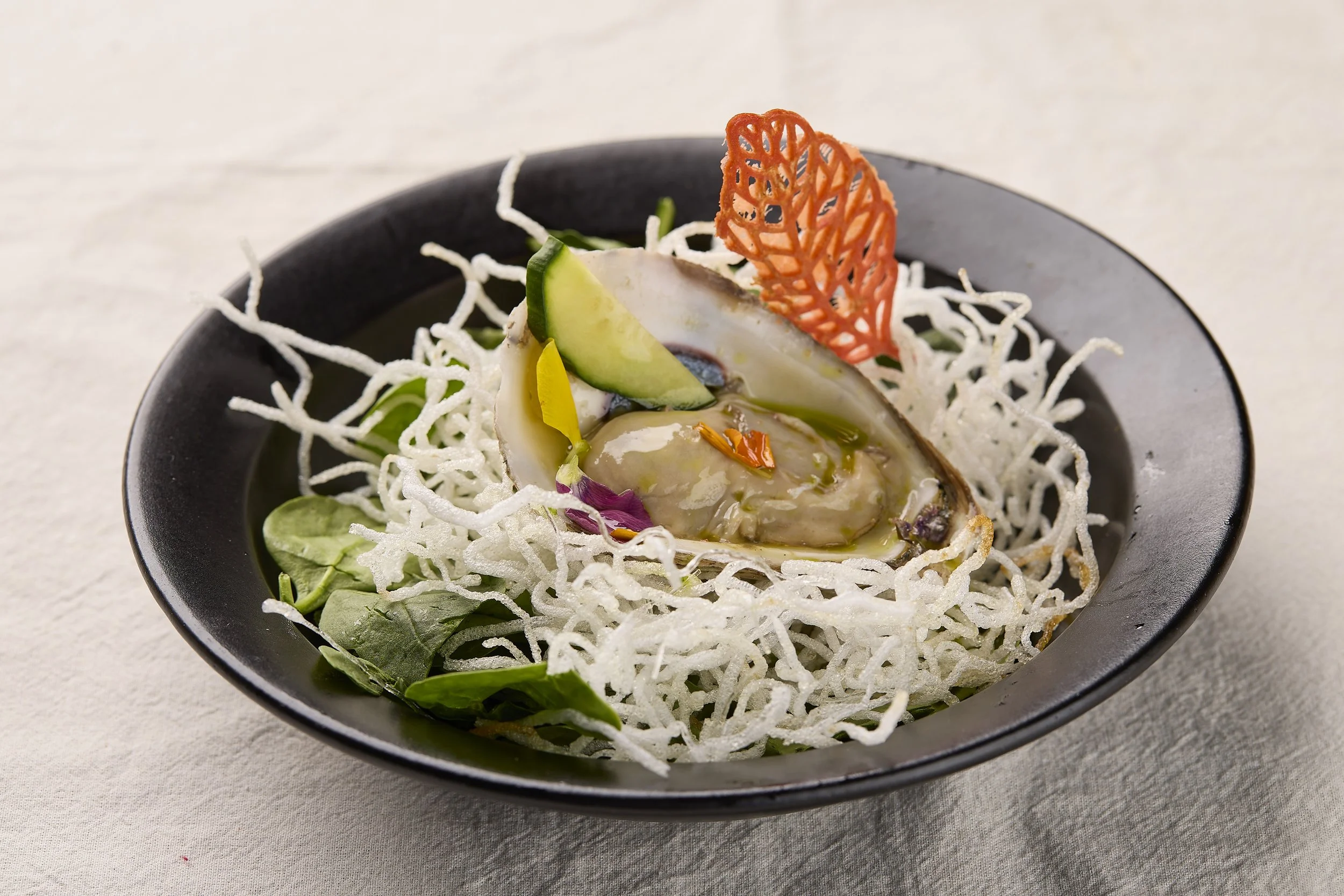 A black bowl containing an oyster served on a bed of shredded white radish, garnished with cucumber slices, edible flowers, and a decorative orange crisp.