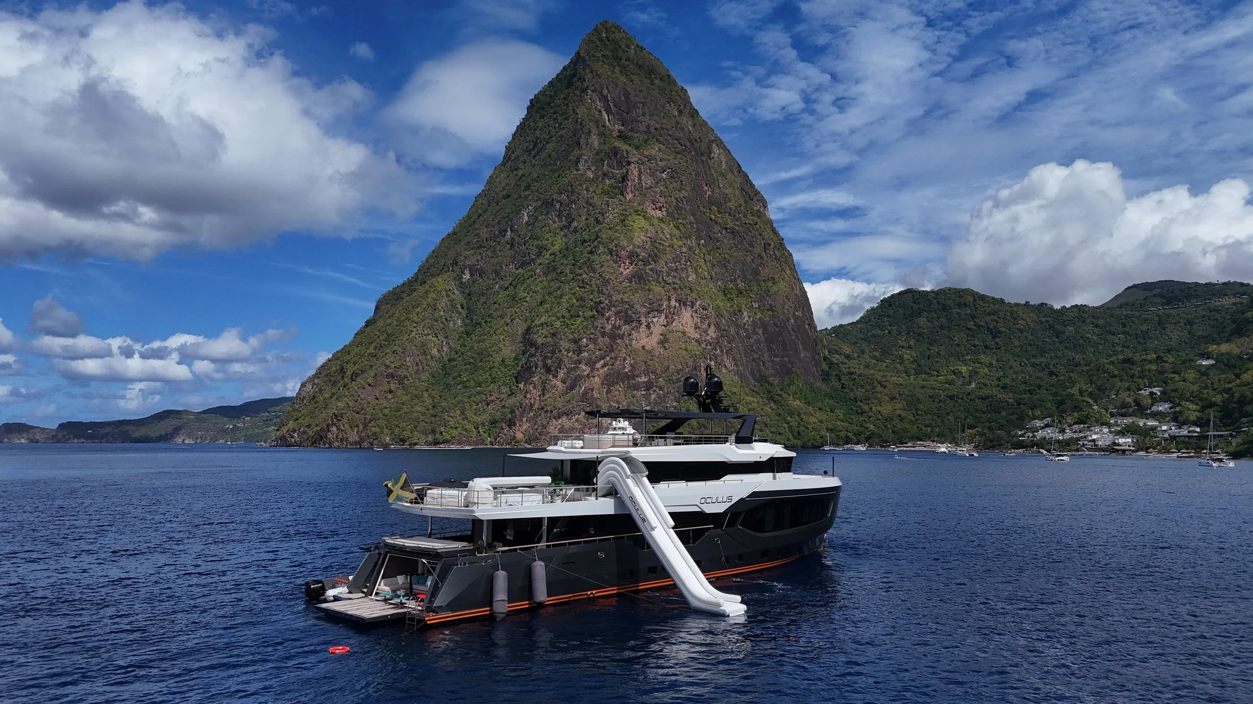 A yacht floating on water near a tall, green-covered mountain with a partly cloudy sky in the background.