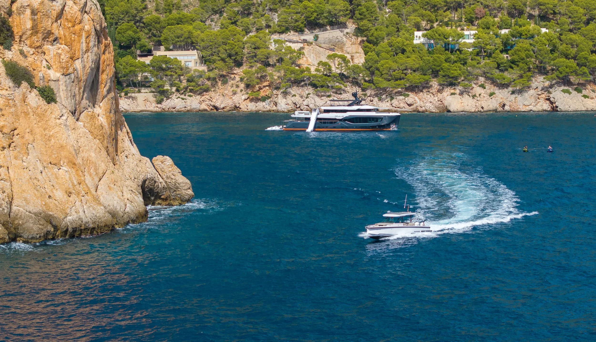 A large boat sailing on blue water near a rocky shoreline with greenery and houses on the hillside.