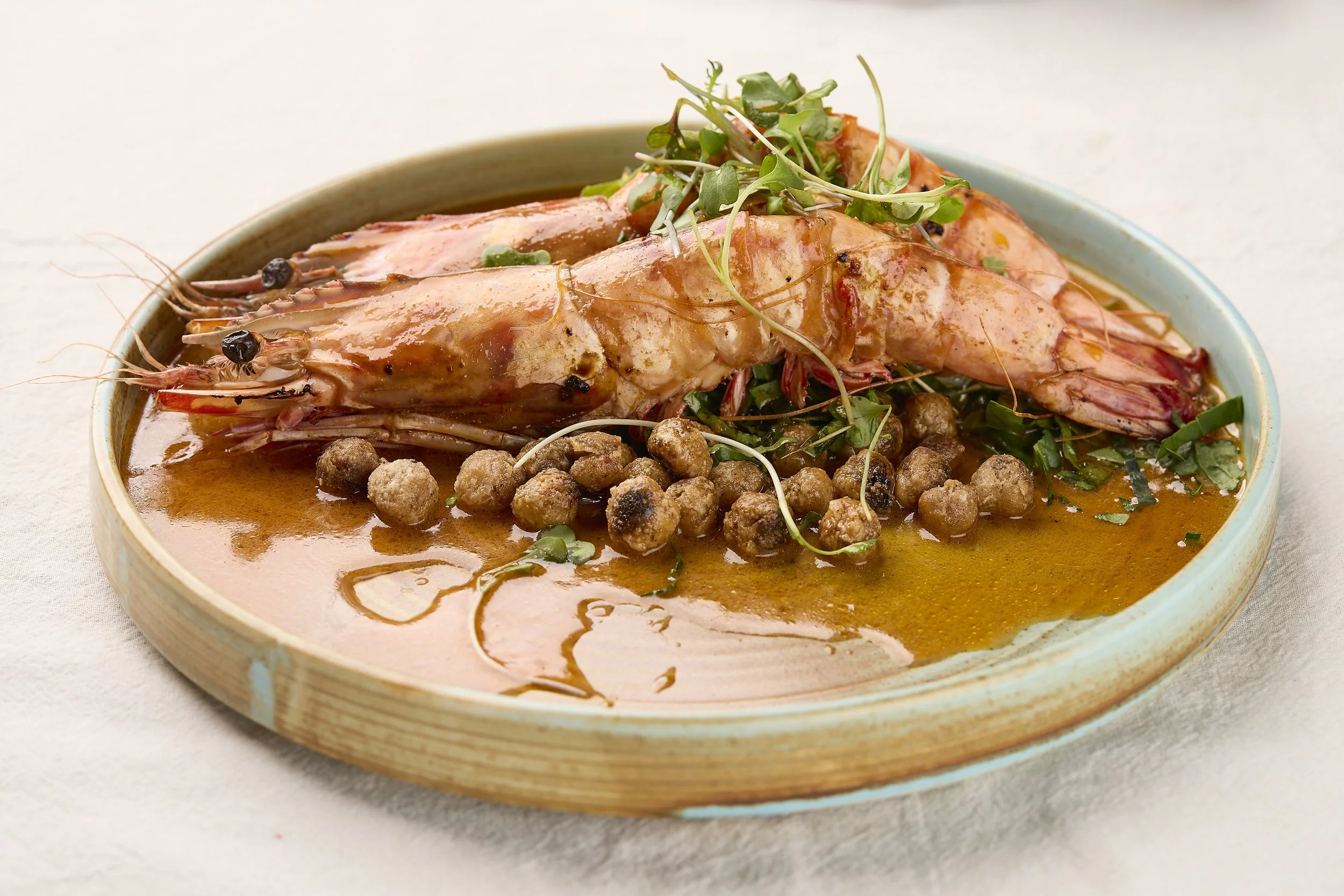 Close-up of a bowl containing cooked shrimp, garnished with microgreens, with a sauce and small meatballs or similar items surrounding the seafood.