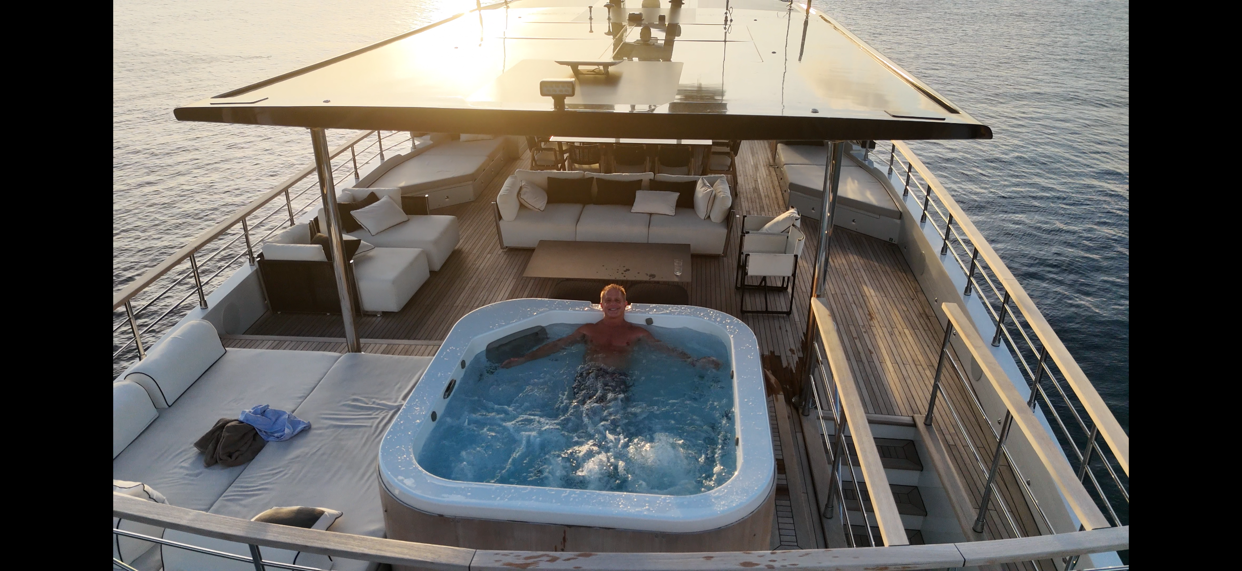 A man relaxing in a hot tub on the deck of a yacht, with outdoor seating and the ocean surrounding the vessel during sunset.