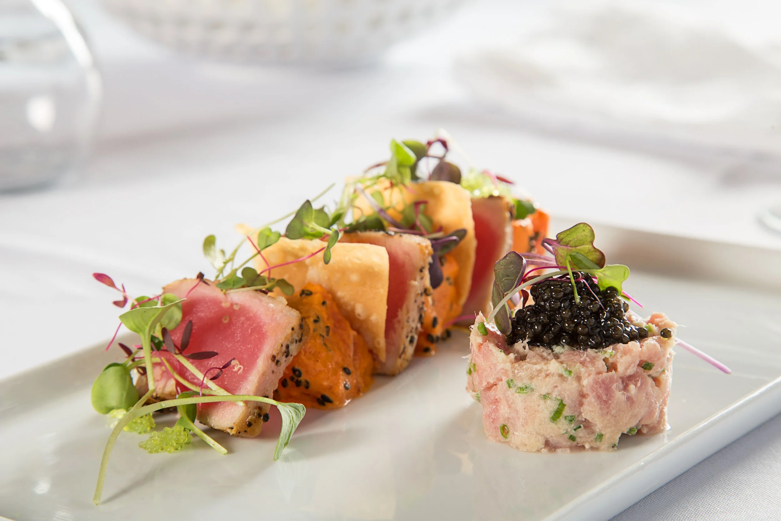 Close-up of an artfully arranged seafood dish with slices of fish or sashimi, garnished with microgreens and a small serving of caviar on a white plate.