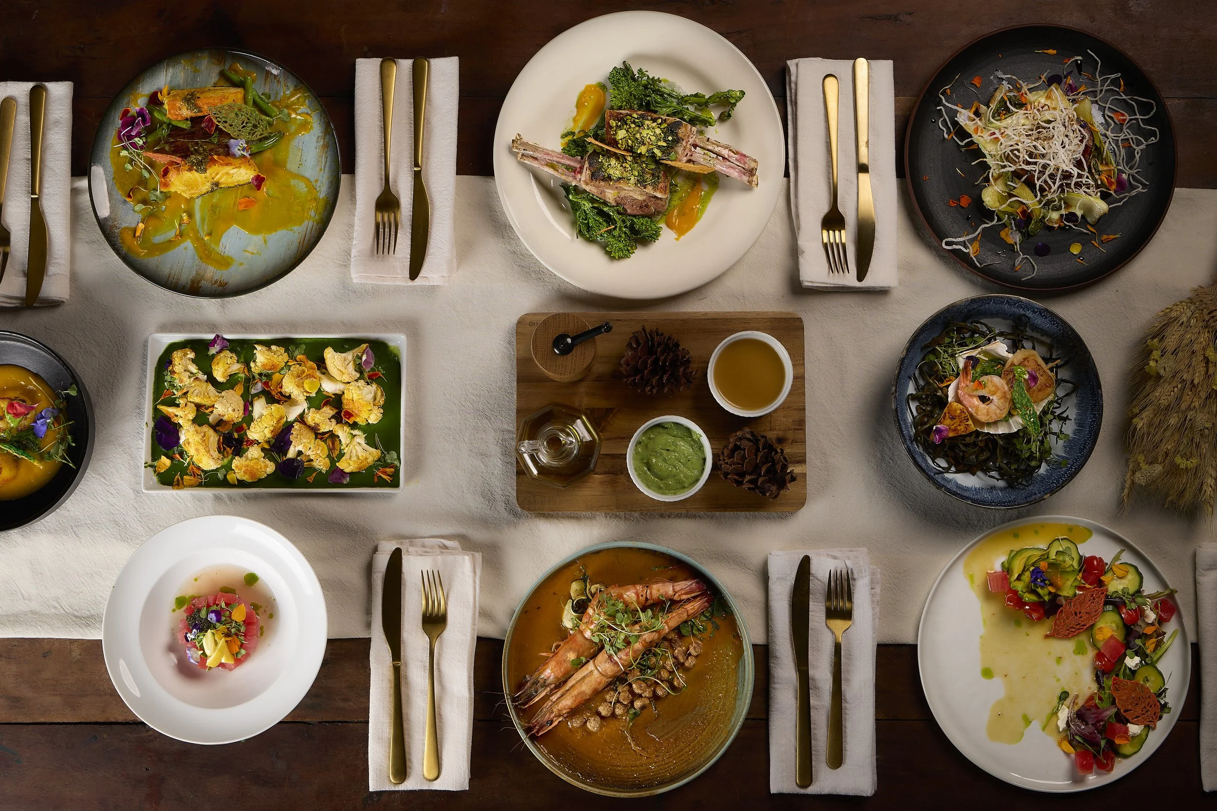 A flat-lay view of multiple plates of gourmet dishes, including a meat dish with green sauce, a prawn dish with vegetables, a colorful soup, and various salads, arranged on a wooden table with gold cutlery.