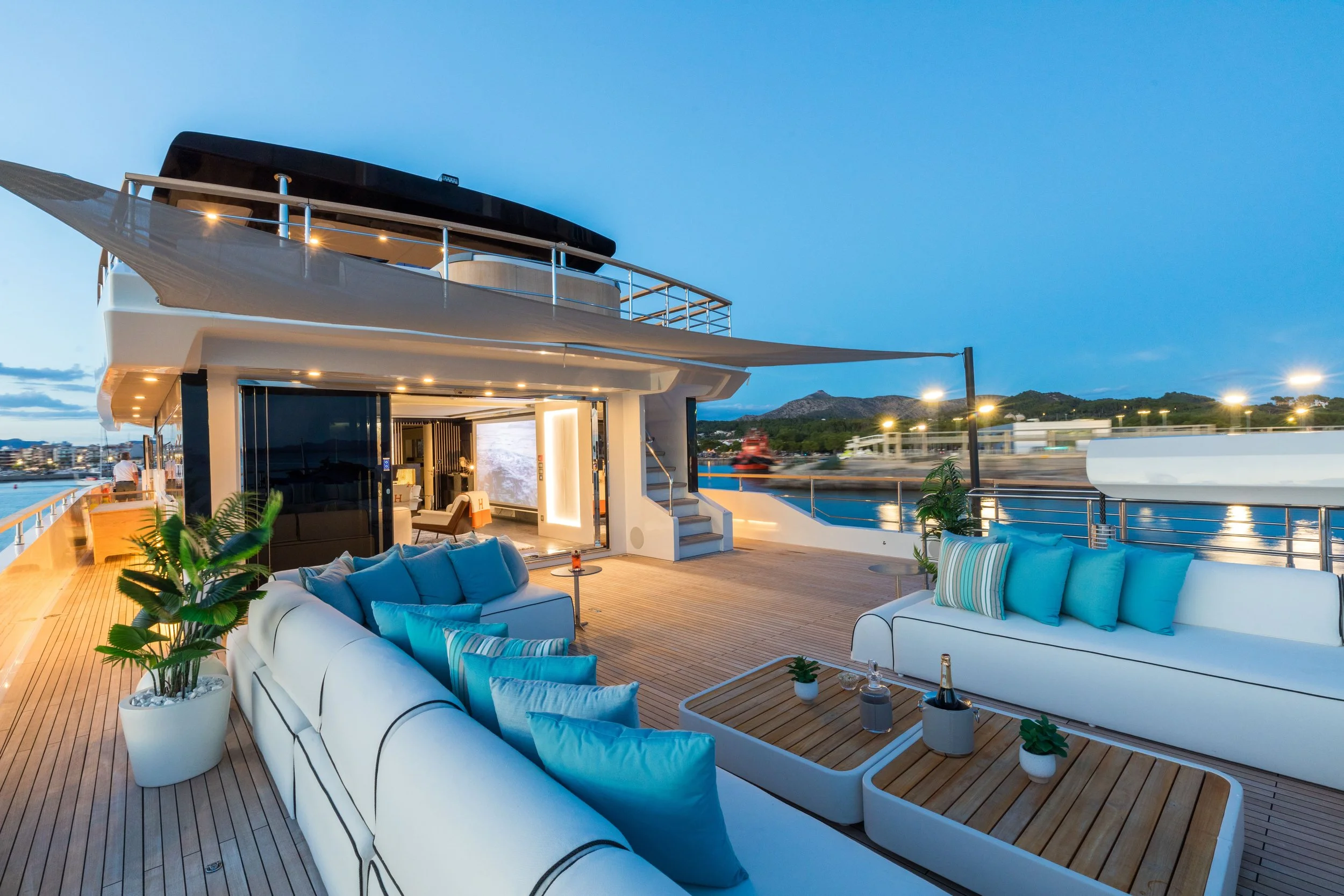 Luxury yacht outdoor lounge area with white sofas, blue pillows, and potted plants, overlooking a marina with lit boats and a mountain in the background during dusk.