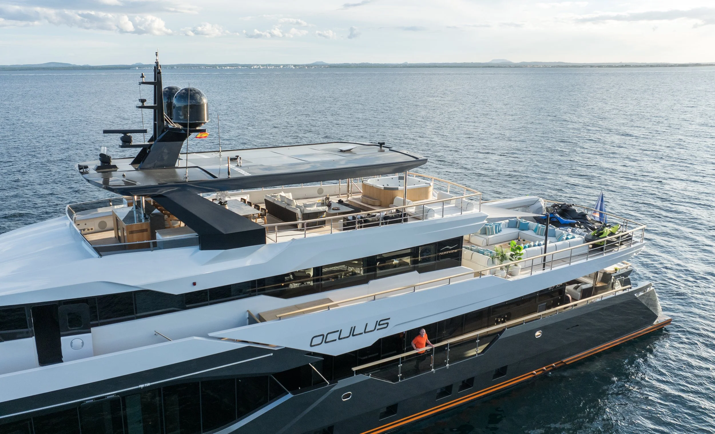 A luxury yacht with outdoor seating, a hot tub, and technological equipment on a calm body of water with a distant shoreline and cloudy sky.