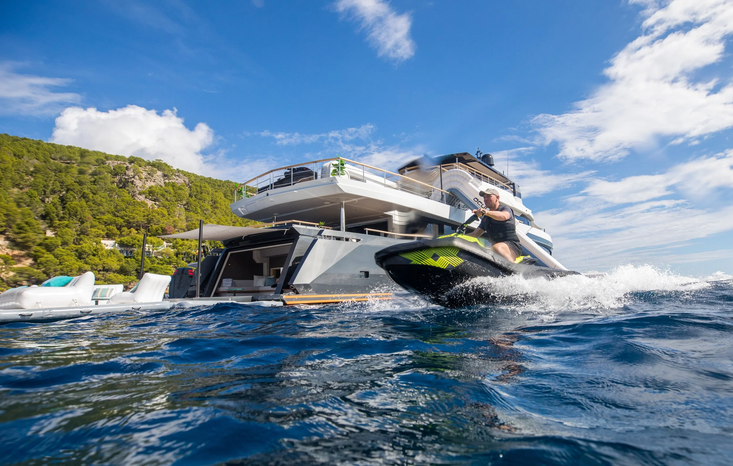 A man is driving a jet ski on the water near a large yacht with a hilly, green landscape in the background under a partly cloudy sky.