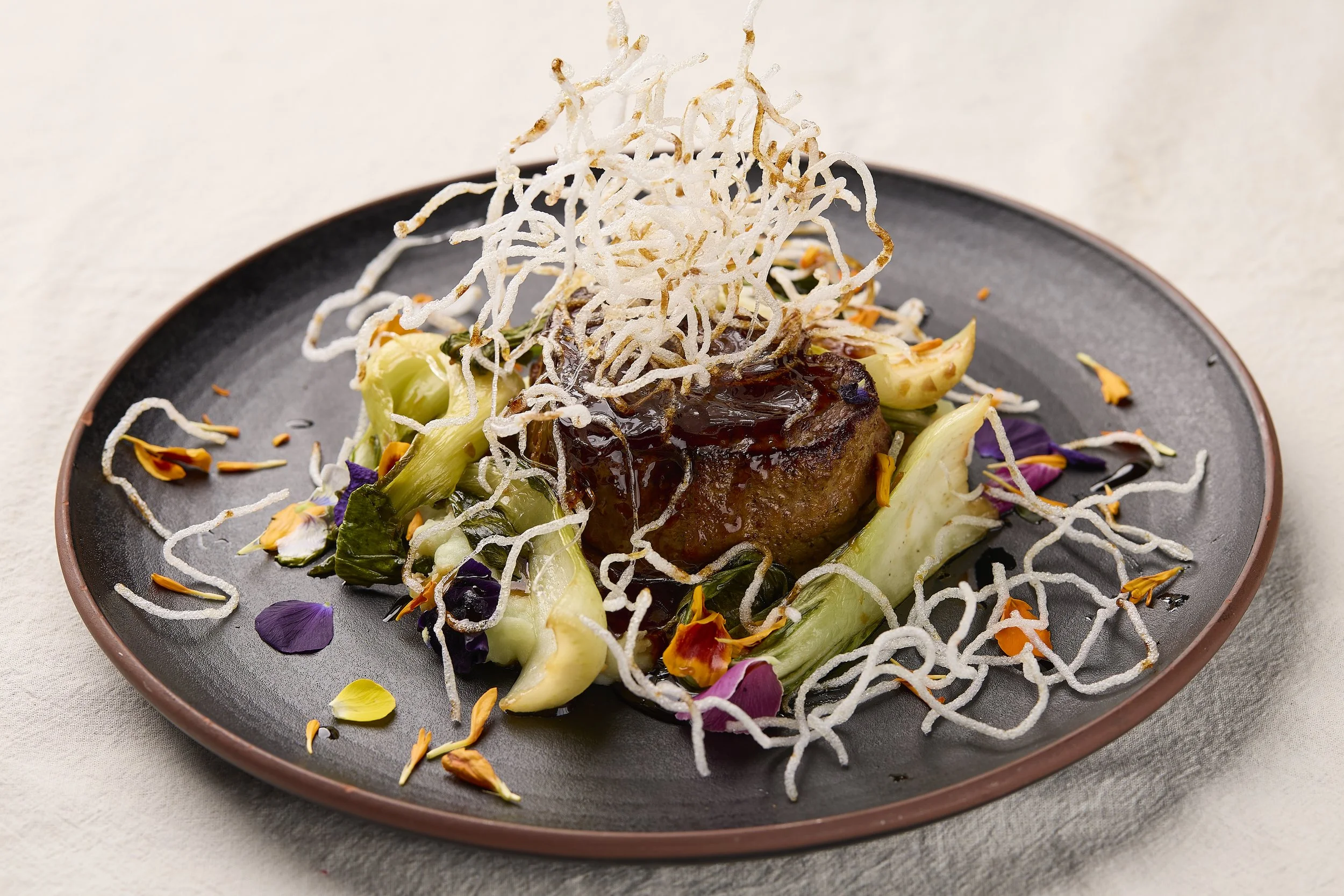 A plated gourmet dish featuring a piece of meat with dark sauce, surrounded by vegetables and garnished with colorful edible flower petals and crispy white wispy toppings.