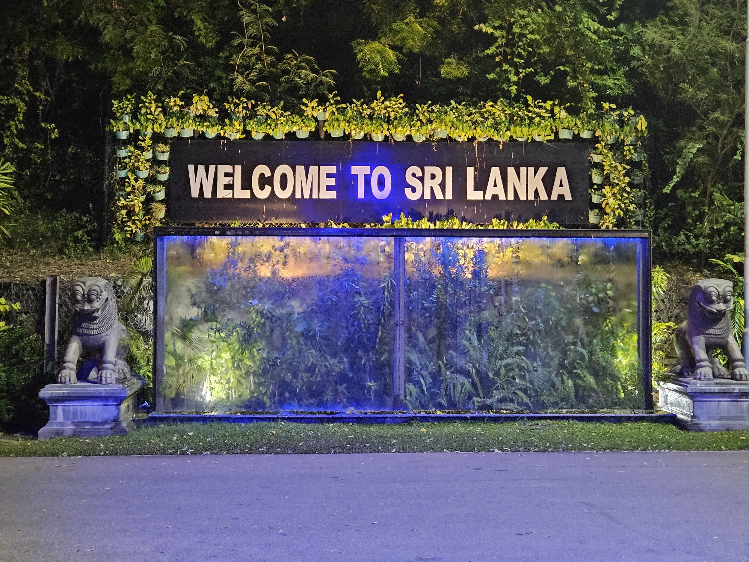Welcoming sign that says 'Welcome to Sri Lanka' with two lion statues on either side, surrounded by lush greenery and illuminated by blue and yellow lights.