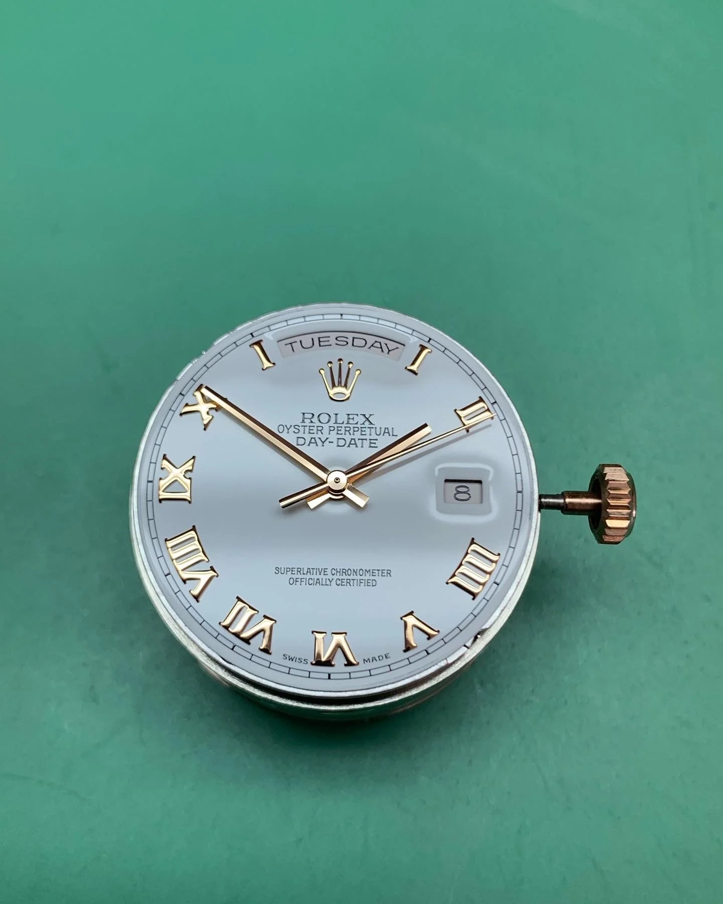 Close-up of a Rolex Oyster Perpetual Day-Date watch with a silver face, gold Roman numeral hour markers, and a date window at 3 o'clock on a green background.