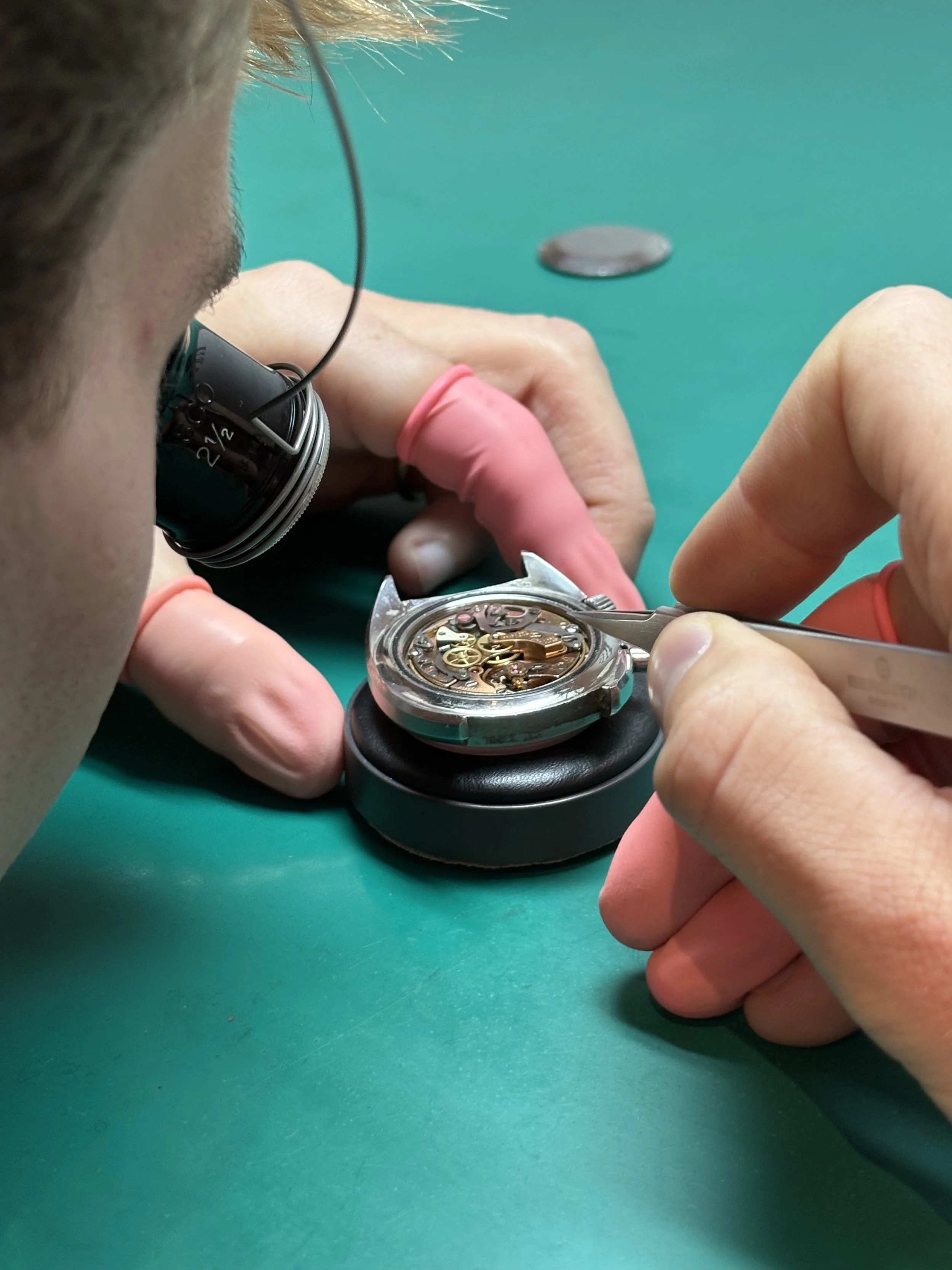 A person wearing pink gloves repairs a mechanical watch using a small tool, with the watch resting on a black cushion on a green work surface.