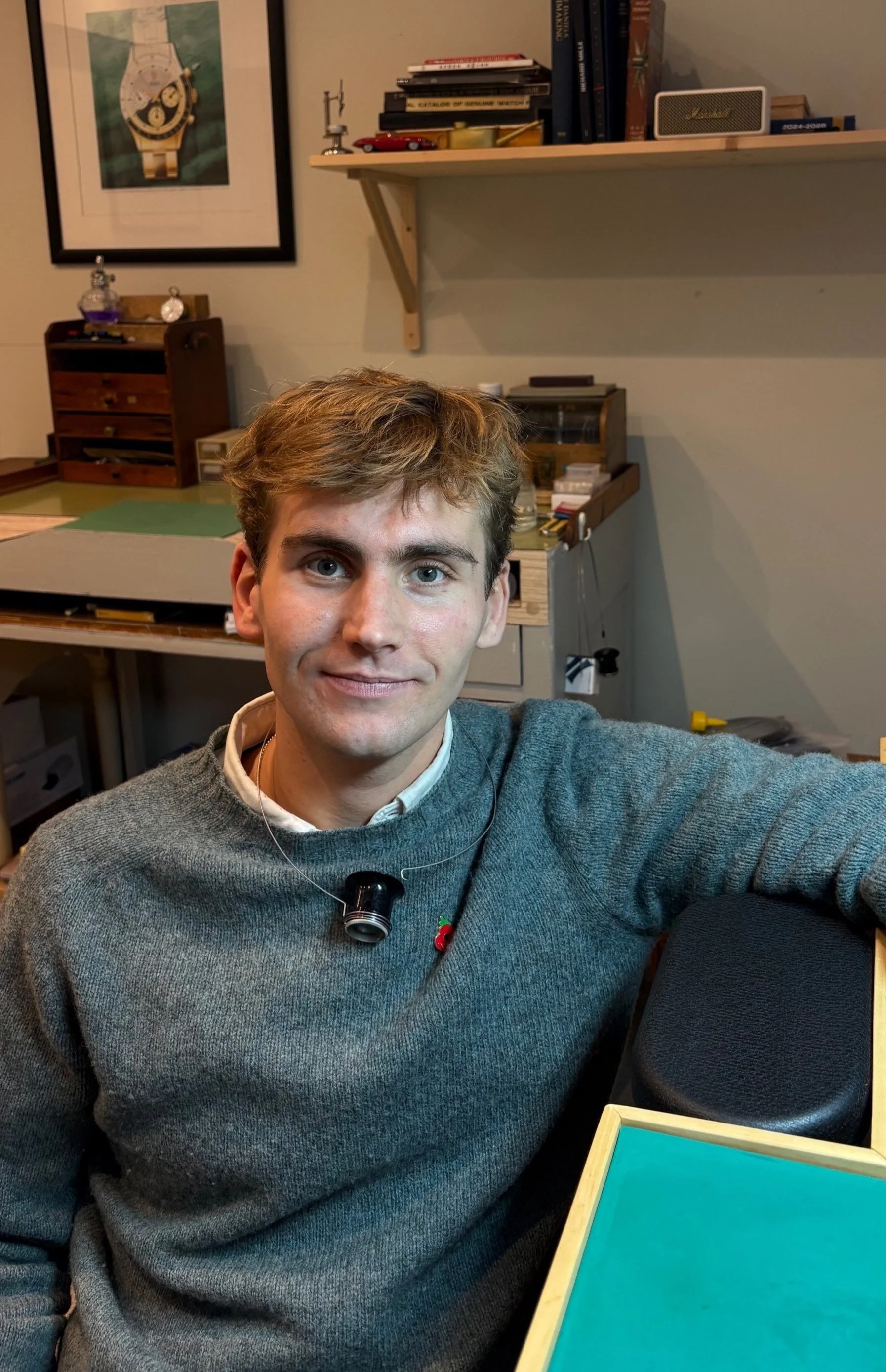 A young man with light brown hair and blue eyes taking a selfie in a room with shelves, books, and artwork in the background.