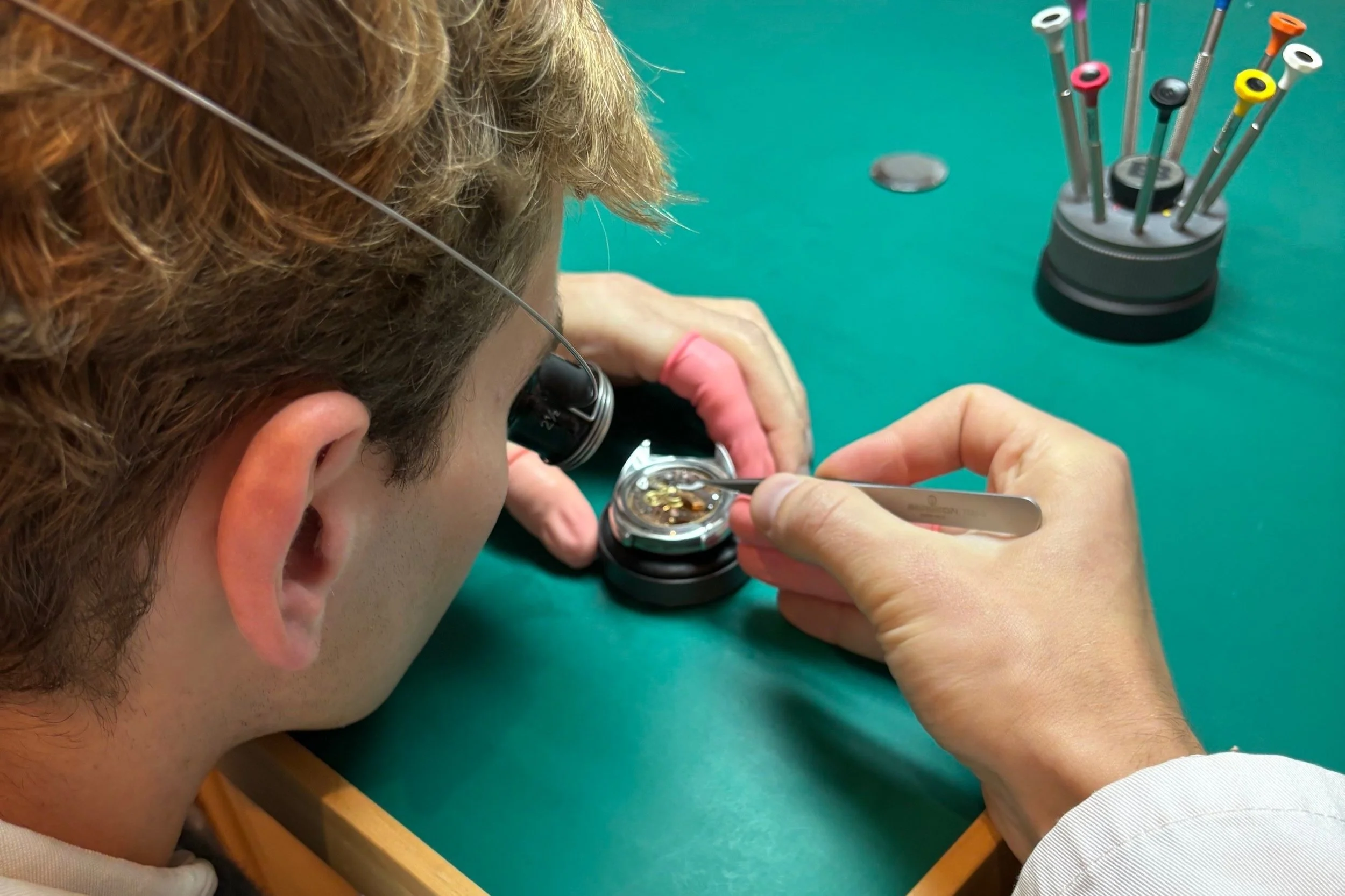 A person with magnifying glasses is working on a small watch component at a watch repair shop, with a set of watch tools in the background.
