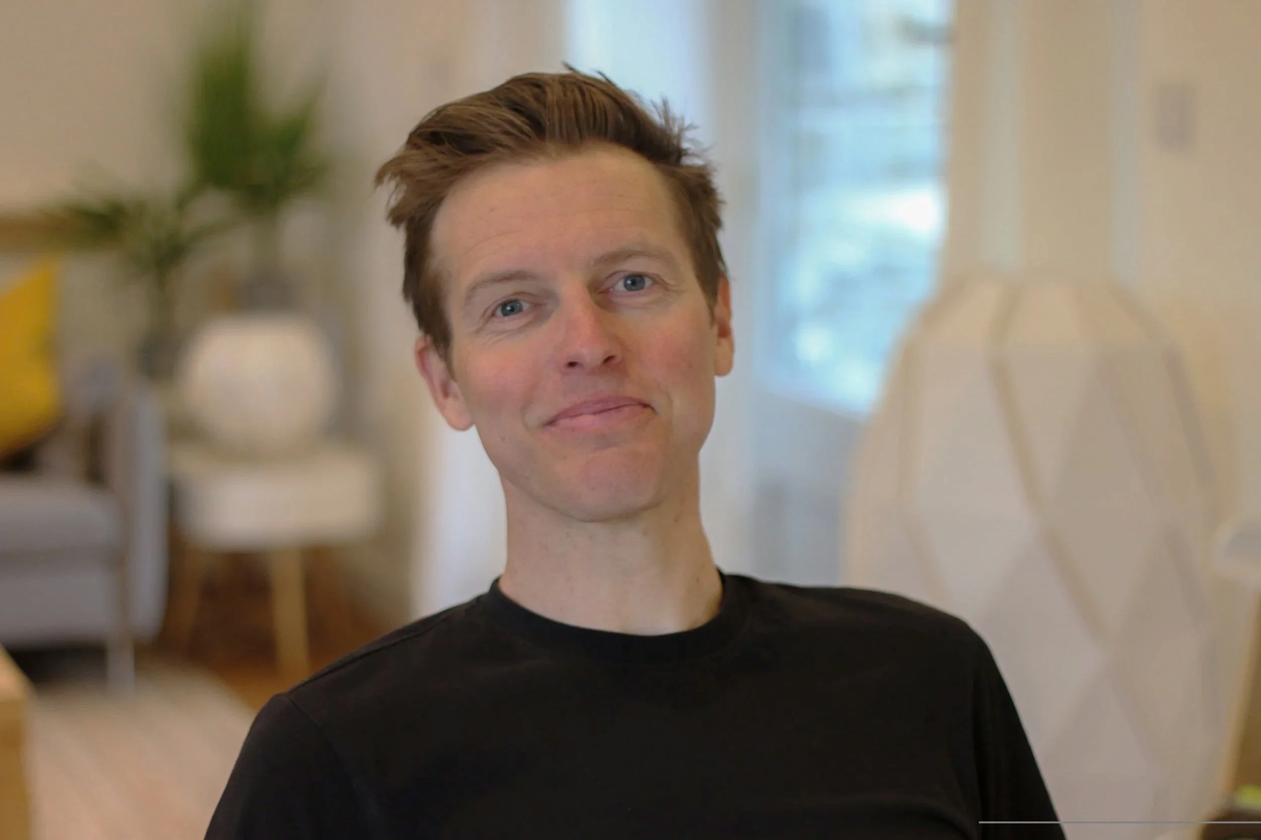 A young man with short red hair and blue eyes, wearing a black t-shirt, smiling softly in a cozy, well-lit room with a blurred background that includes a plant, bookshelf, and window.