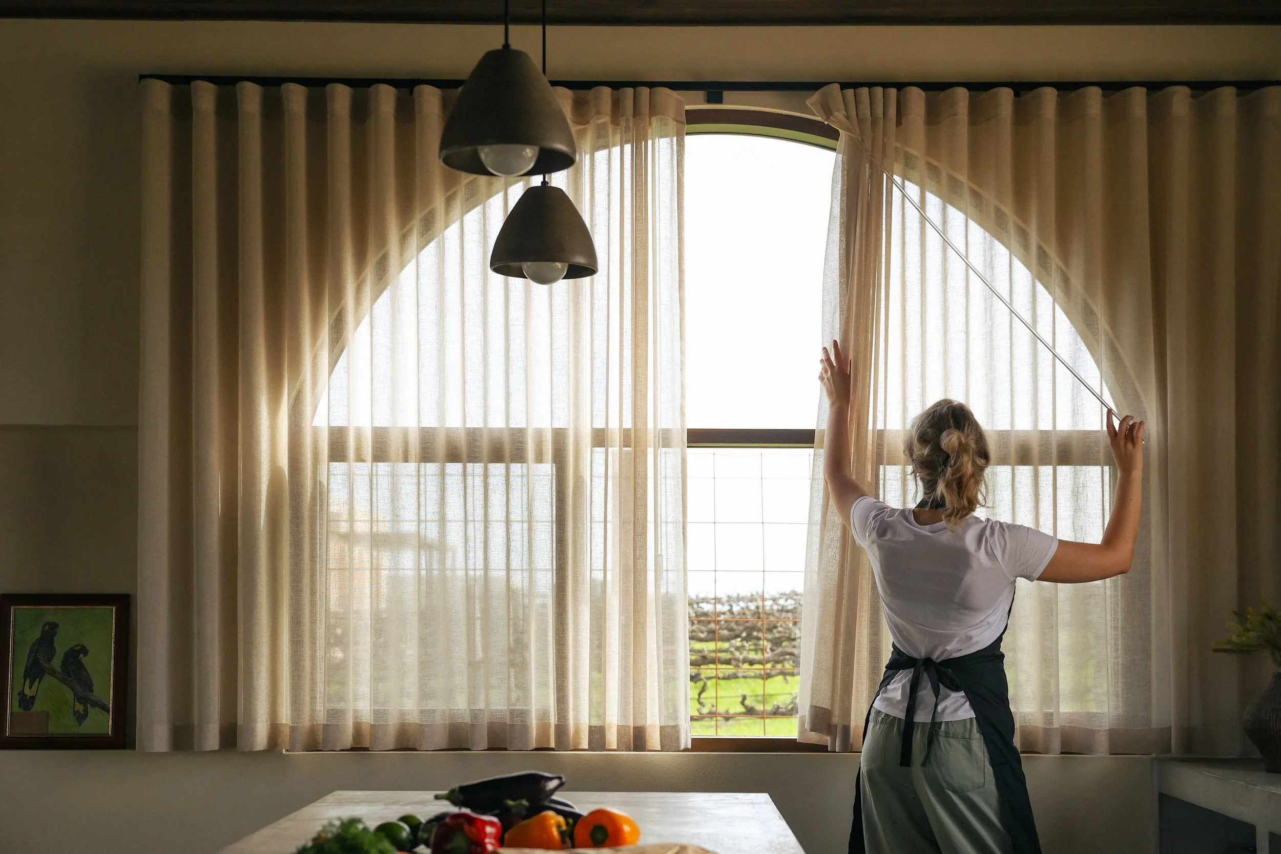 A woman standing by a window, pulling back a beige curtain with her left hand and holding tie-back cord with her right hand, in a kitchen with a table and colorful vegetables.