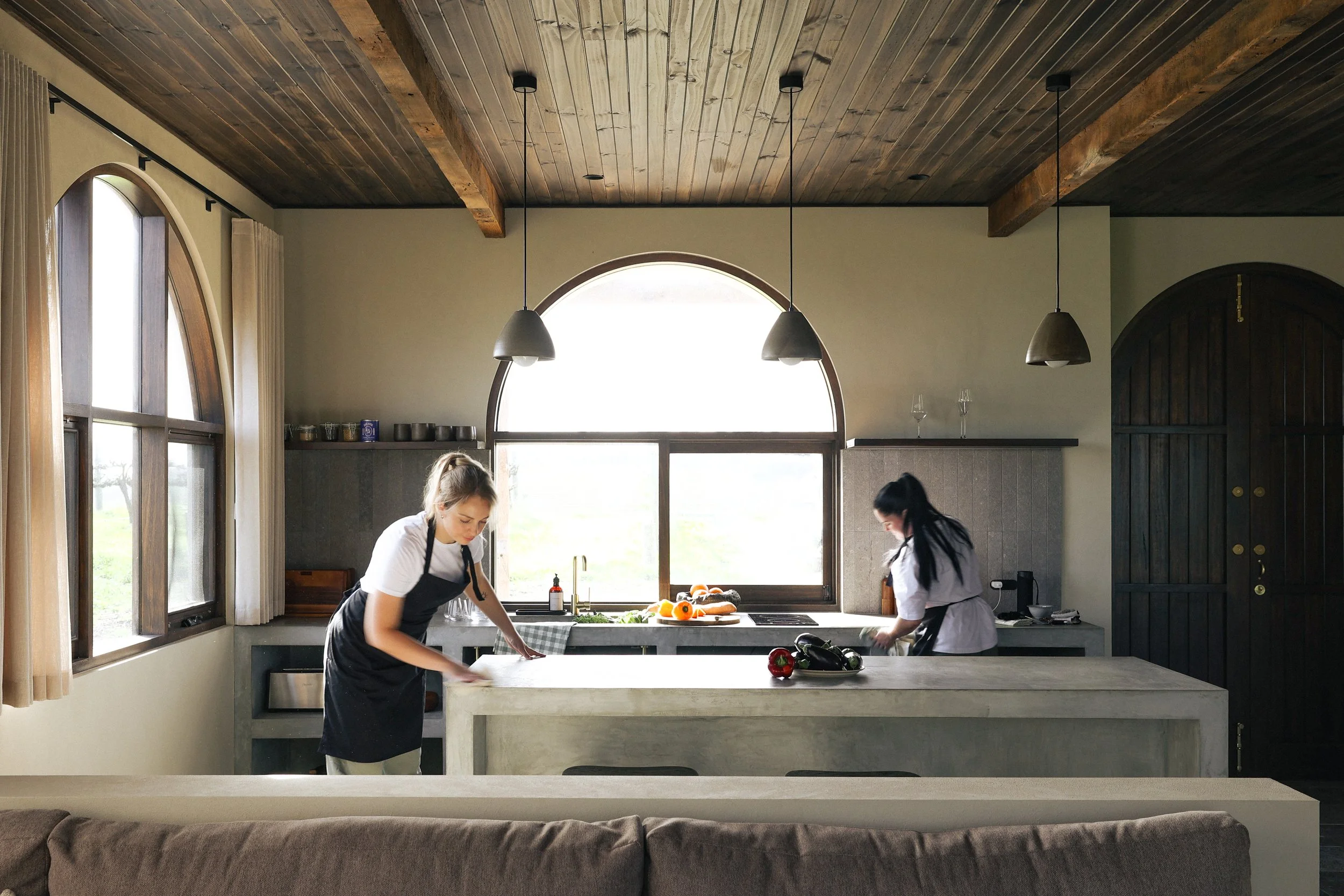 Two women cleaning a kitchen island with vegetables, in a kitchen with large arched windows and a wooden ceiling.