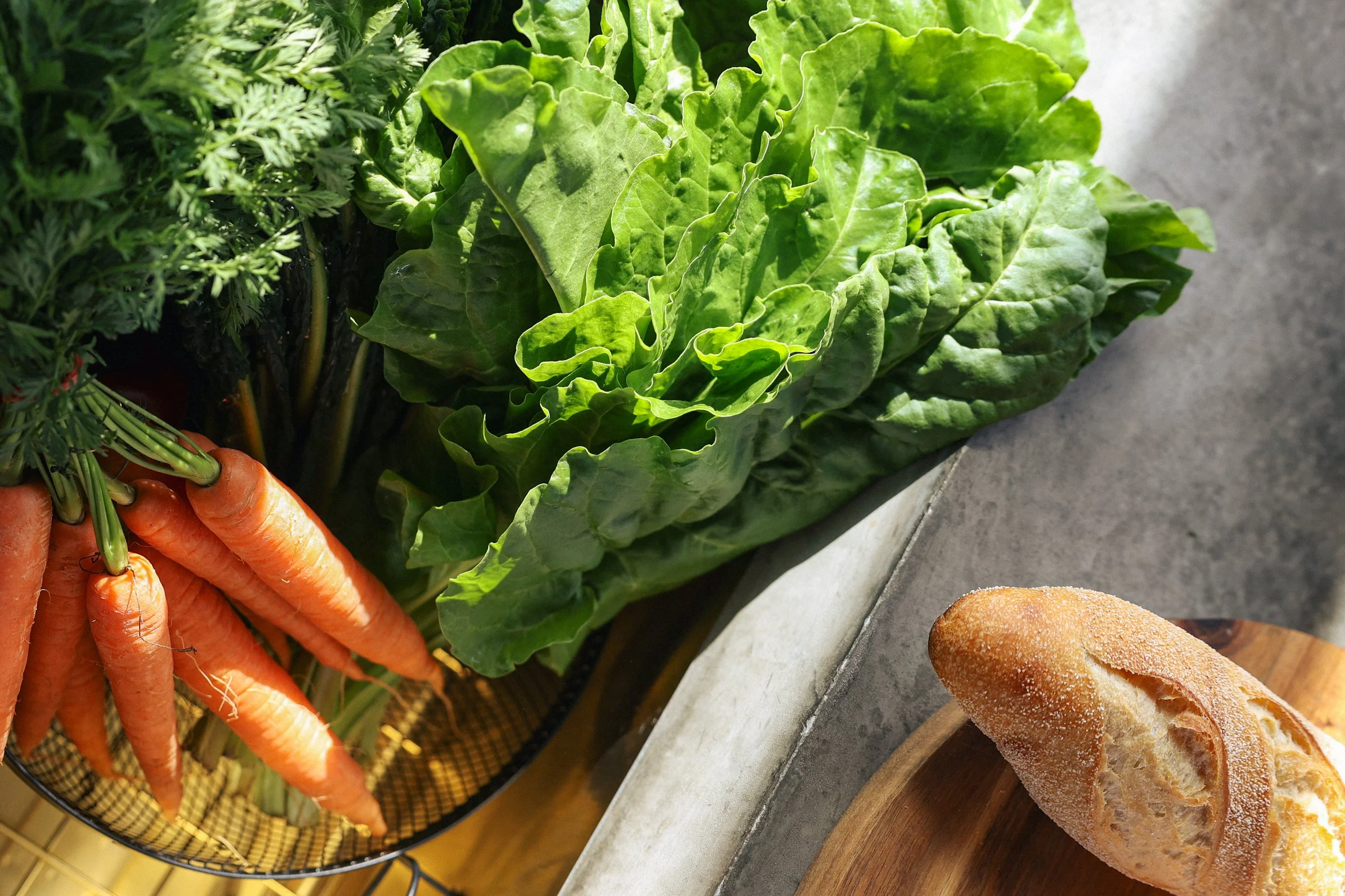 Fresh carrots with green tops, leafy lettuce, and a loaf of bread on a wooden cutting board.