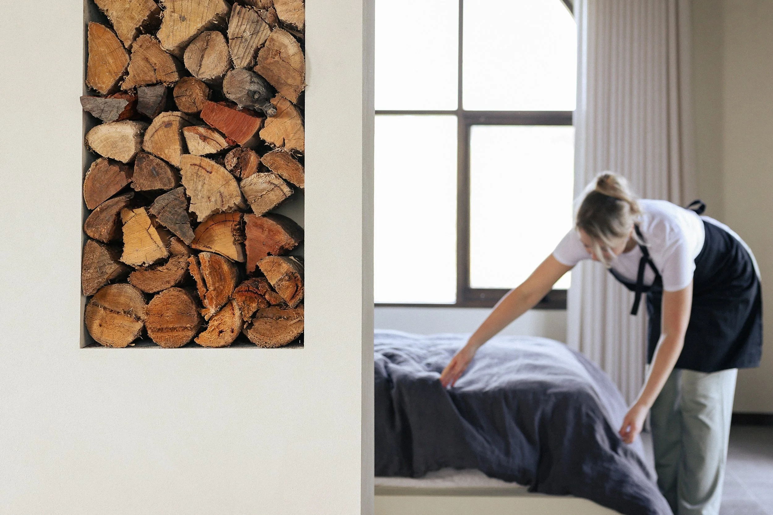 A woman in a black apron tidying a bed in a bedroom with a window, and a section of wall with a built-in nook filled with stacked firewood.