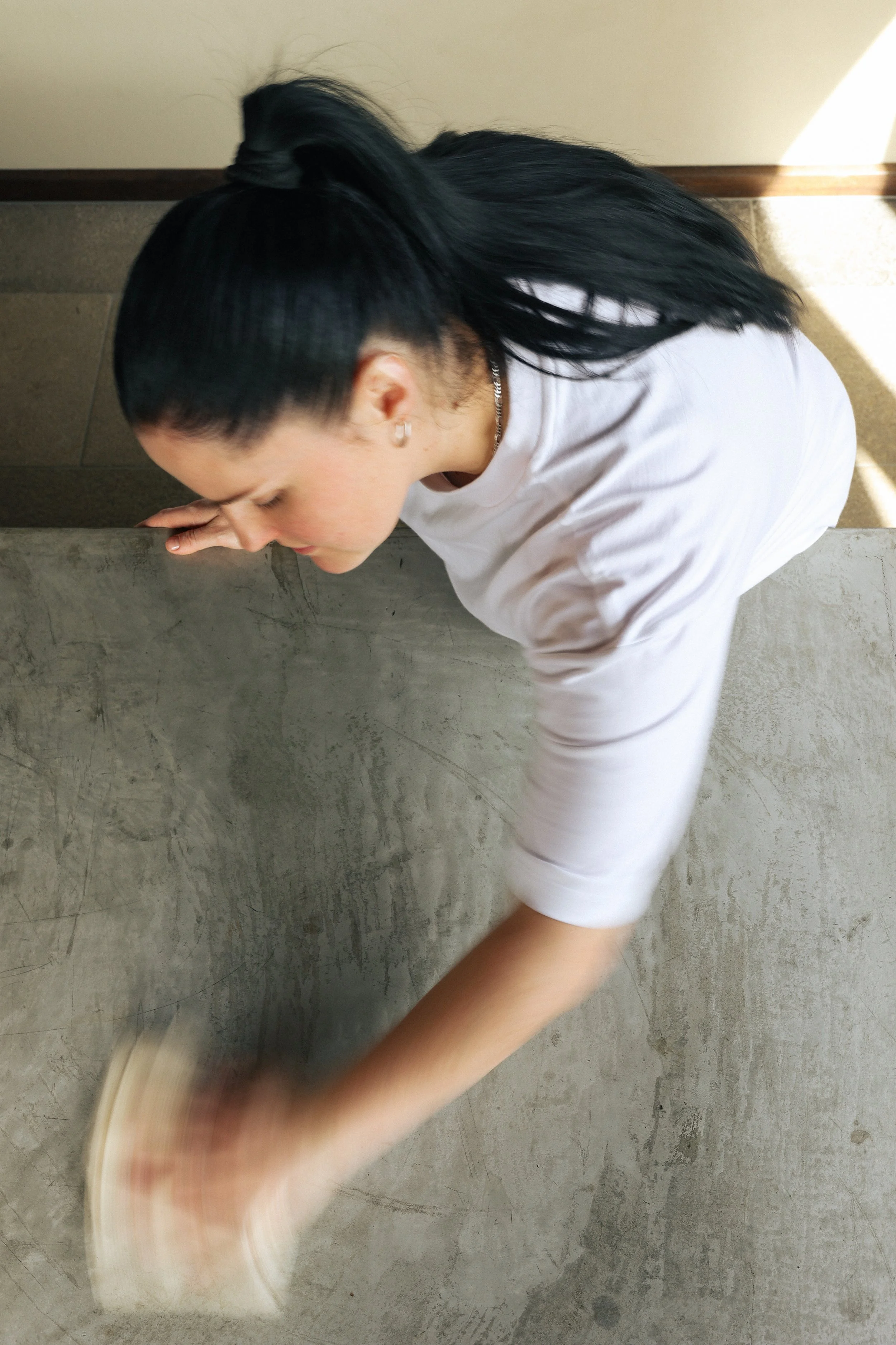 A woman with long black hair tied back, wearing a white t-shirt, wiping or cleaning a gray concrete surface with a cloth or sponge.