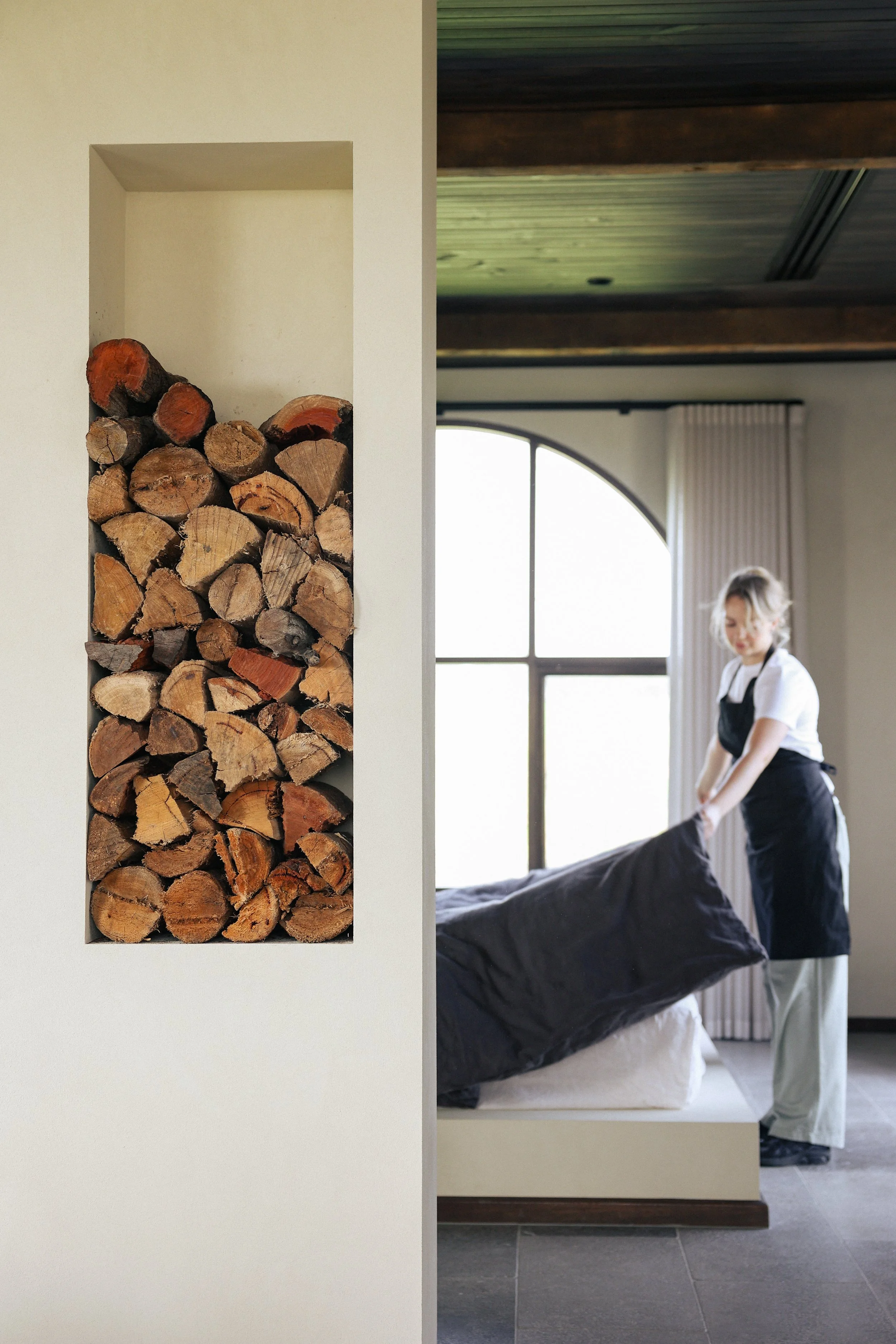 A woman making a bed in a bedroom with a large window and wood ceiling, seen through an opening in the wall with a built-in fireplace filled with firewood.