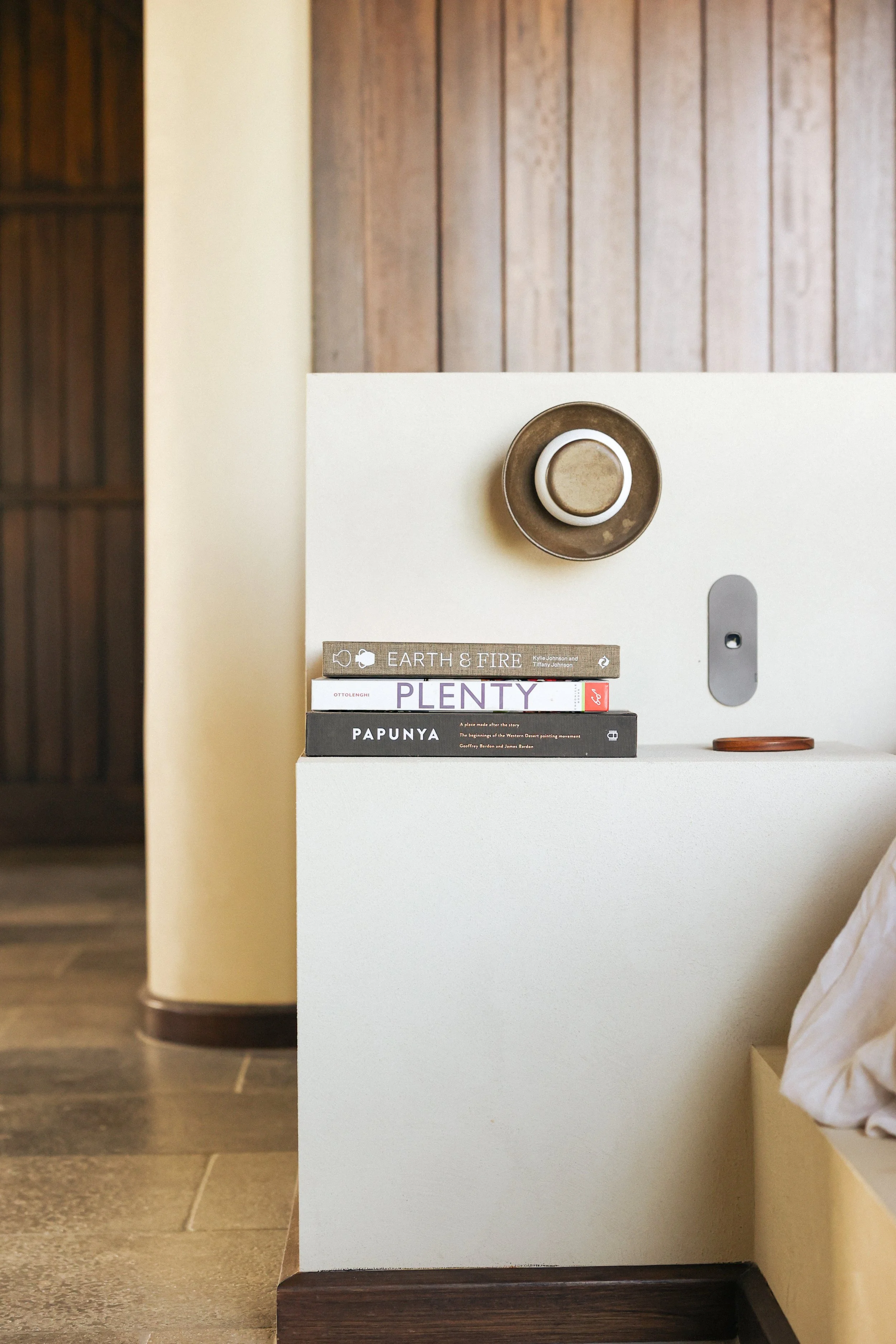 A minimalist bedroom nightstand with three stacked books, a small decorative object, and a round wall feature, with wood-paneled wall and a bed partially visible.