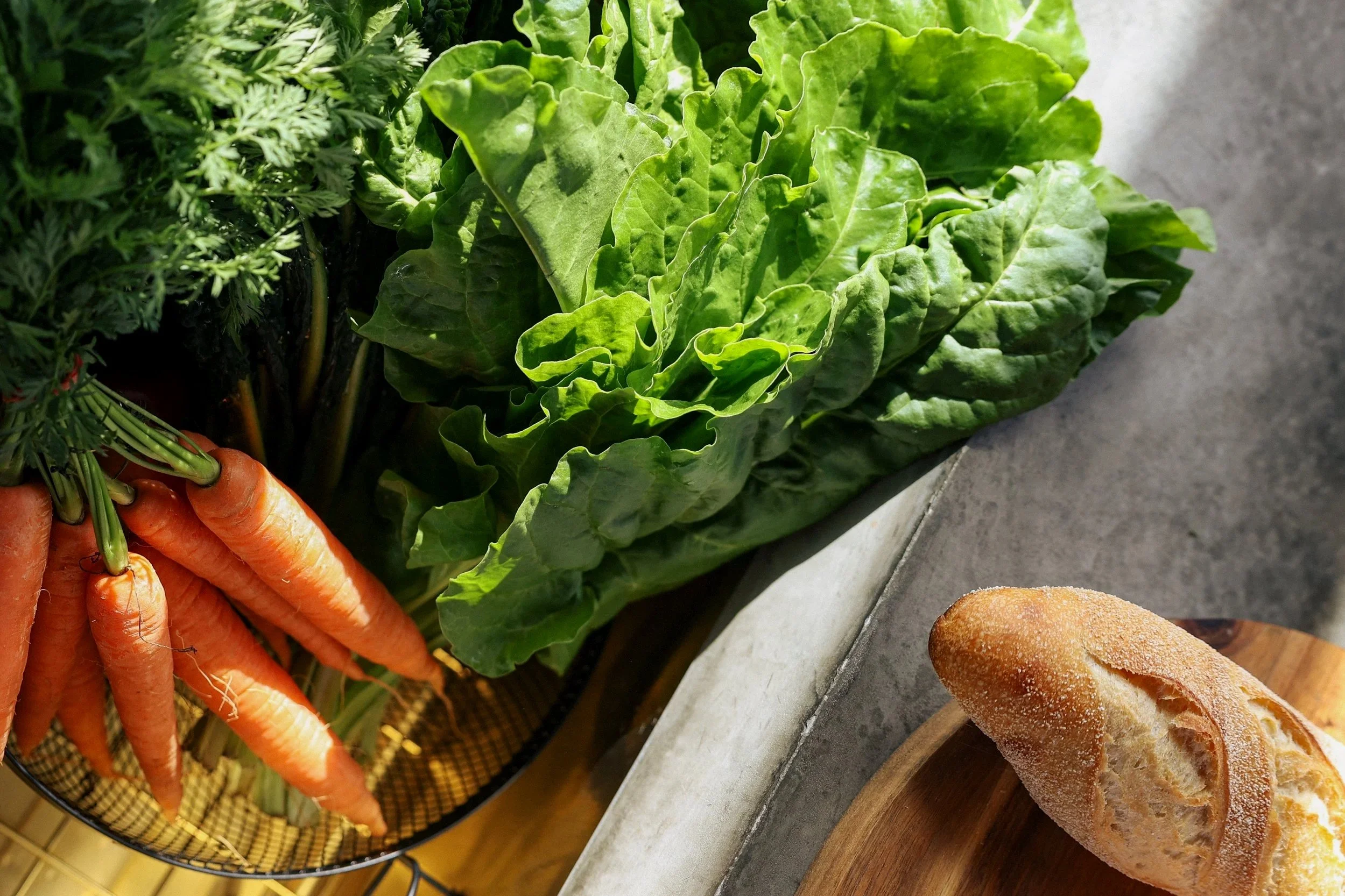 Fresh carrots, leafy lettuce, and greens arranged on a kitchen counter next to a loaf of bread on a wooden cutting board.