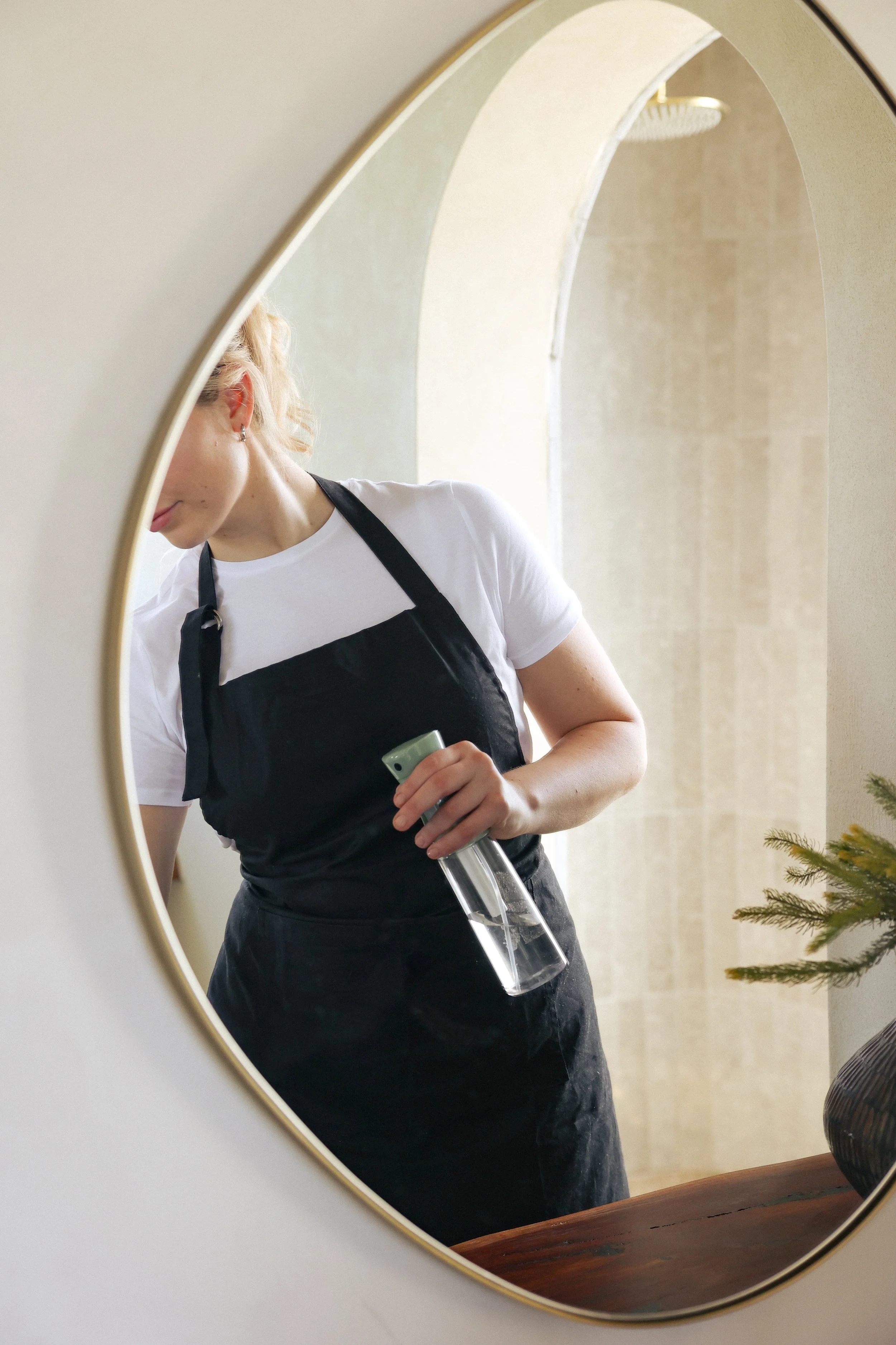 A woman with blonde hair in a ponytail wearing a white t-shirt and black apron, holding a spray bottle, seen through a mirror.