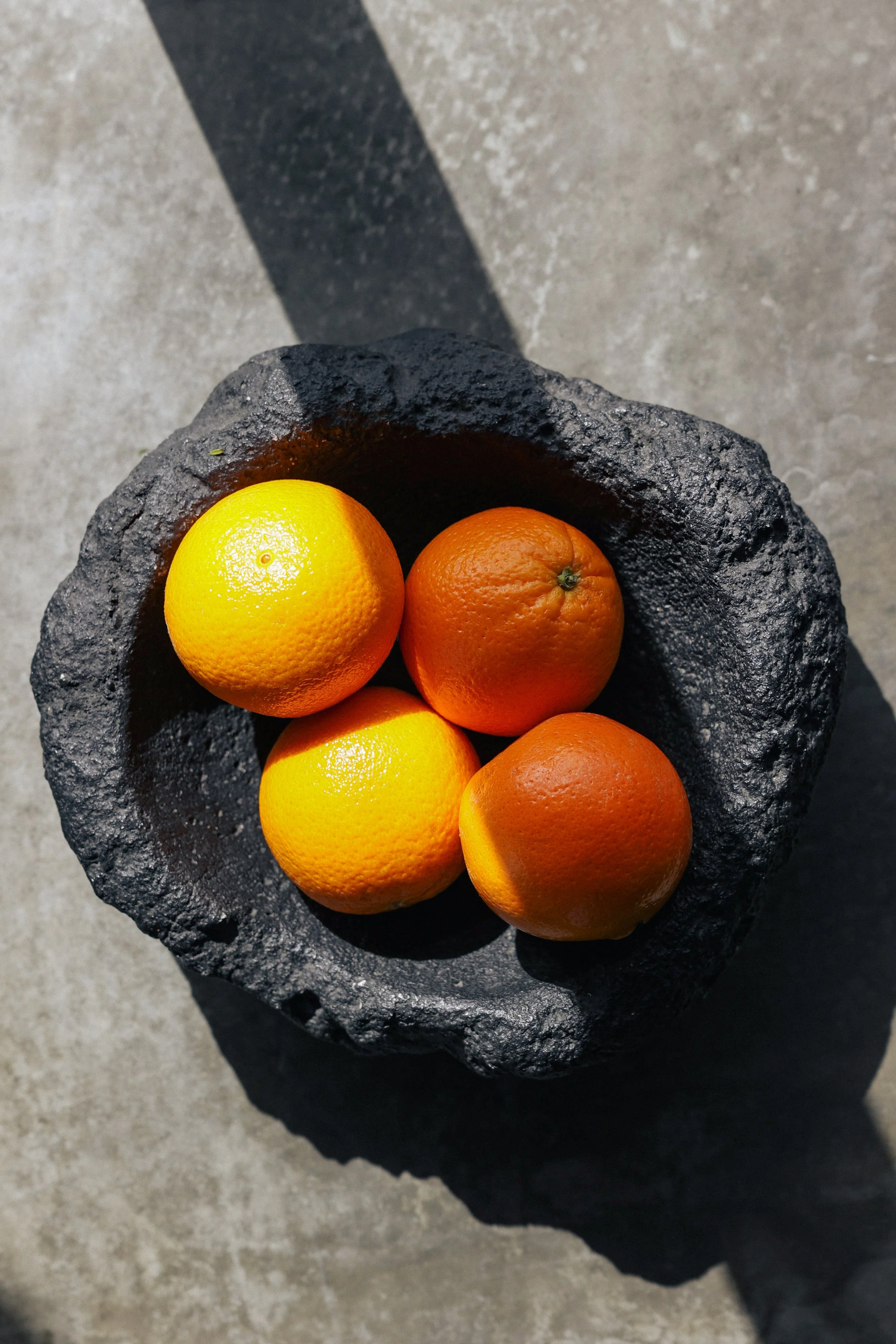 Four oranges in a black stone bowl on a concrete surface with sunlight and shadow.