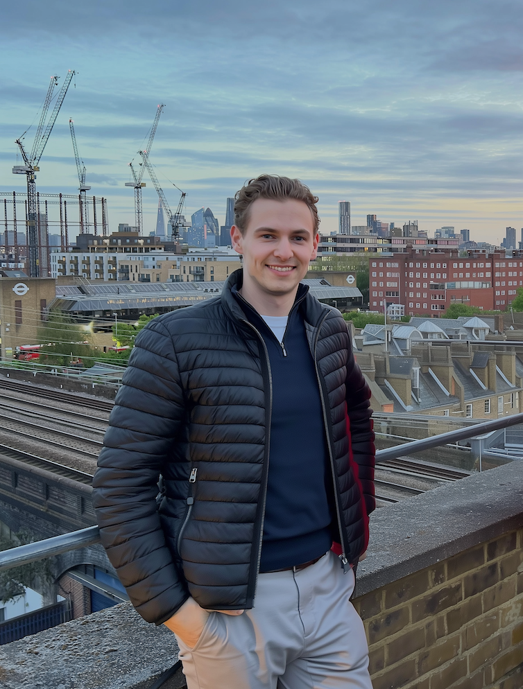 A young man smiling and posing on a rooftop with cityscape, including construction cranes, in the background during the daytime.
