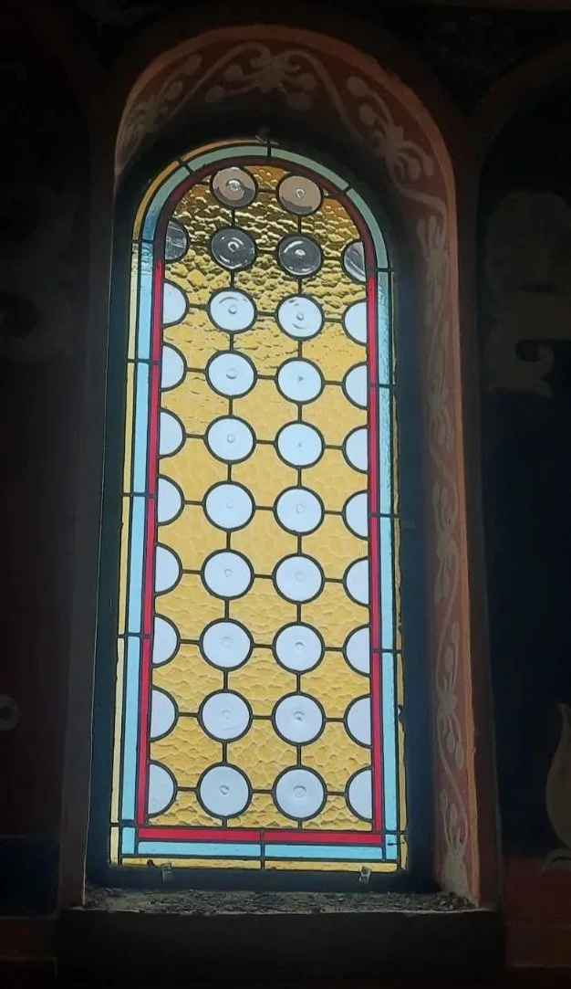 Colorful stained glass window with a yellow background and circular glass pieces arranged in a grid, surrounded by a decorative border.