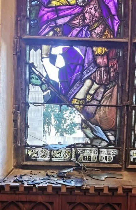 A stained glass window with a wooden frame, featuring colorful purple, yellow, and other stained glass pieces, with some broken glass and debris on the window sill below.