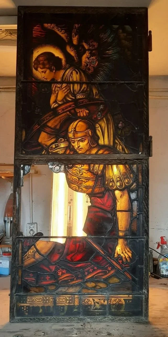 A stained glass window depicting a knight kneeling with a sword, and an angel or saint above him with a halo, set in a dark wooden frame.