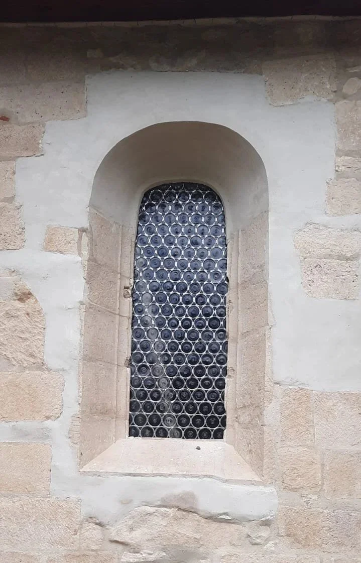 A small arched window in a stone wall filled with empty glass bottles arranged in a grid pattern.