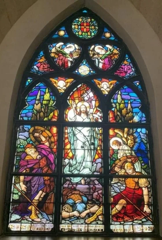 Colorful stained glass window depicting the resurrection of Jesus Christ, with angels, soldiers, and religious symbols.