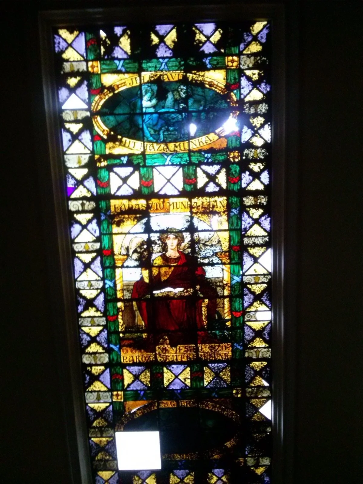 Stained glass window depicting a seated person with a halo, holding a book, with decorative text and colorful geometric patterns.