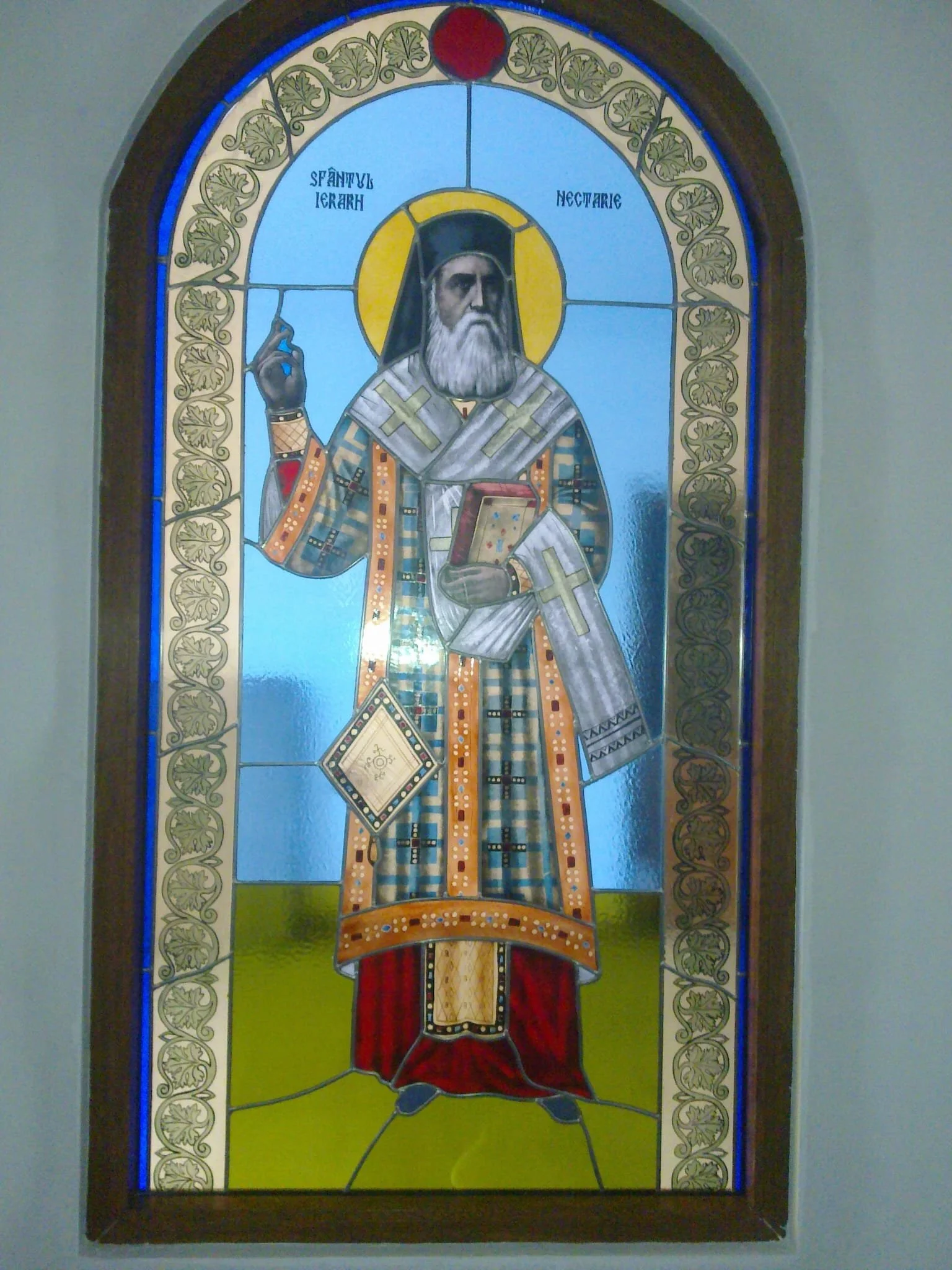 Stained glass depiction of a saint holding a book and making a blessing gesture, featuring various patterns and symbols, with a halo around his head and Greek script in the background.