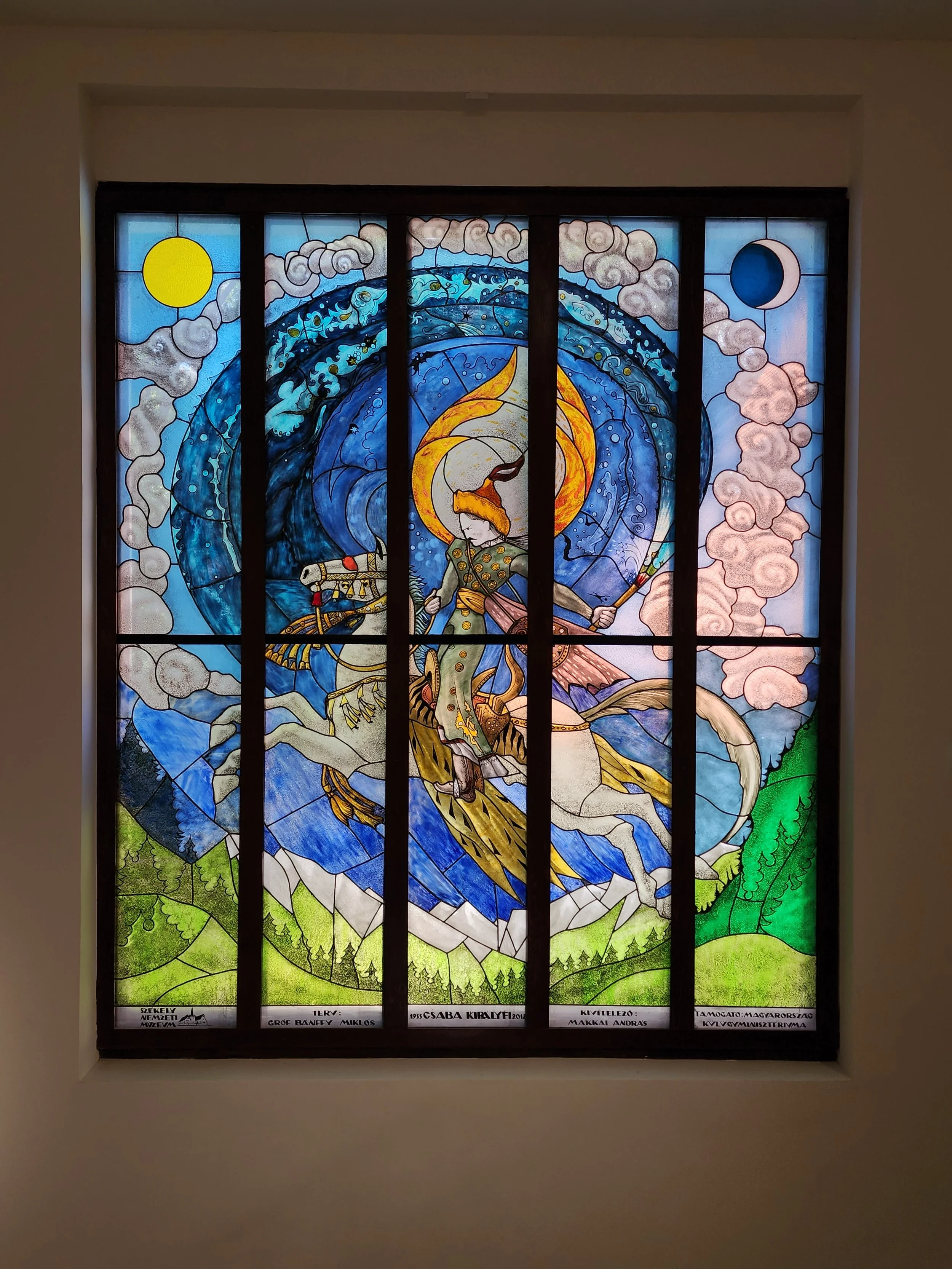 Colorful stained glass window depicting a knight on a white horse with a sword, surrounded by clouds, a blue wave, and a yellow sun on the left and a blue moon on the right.
