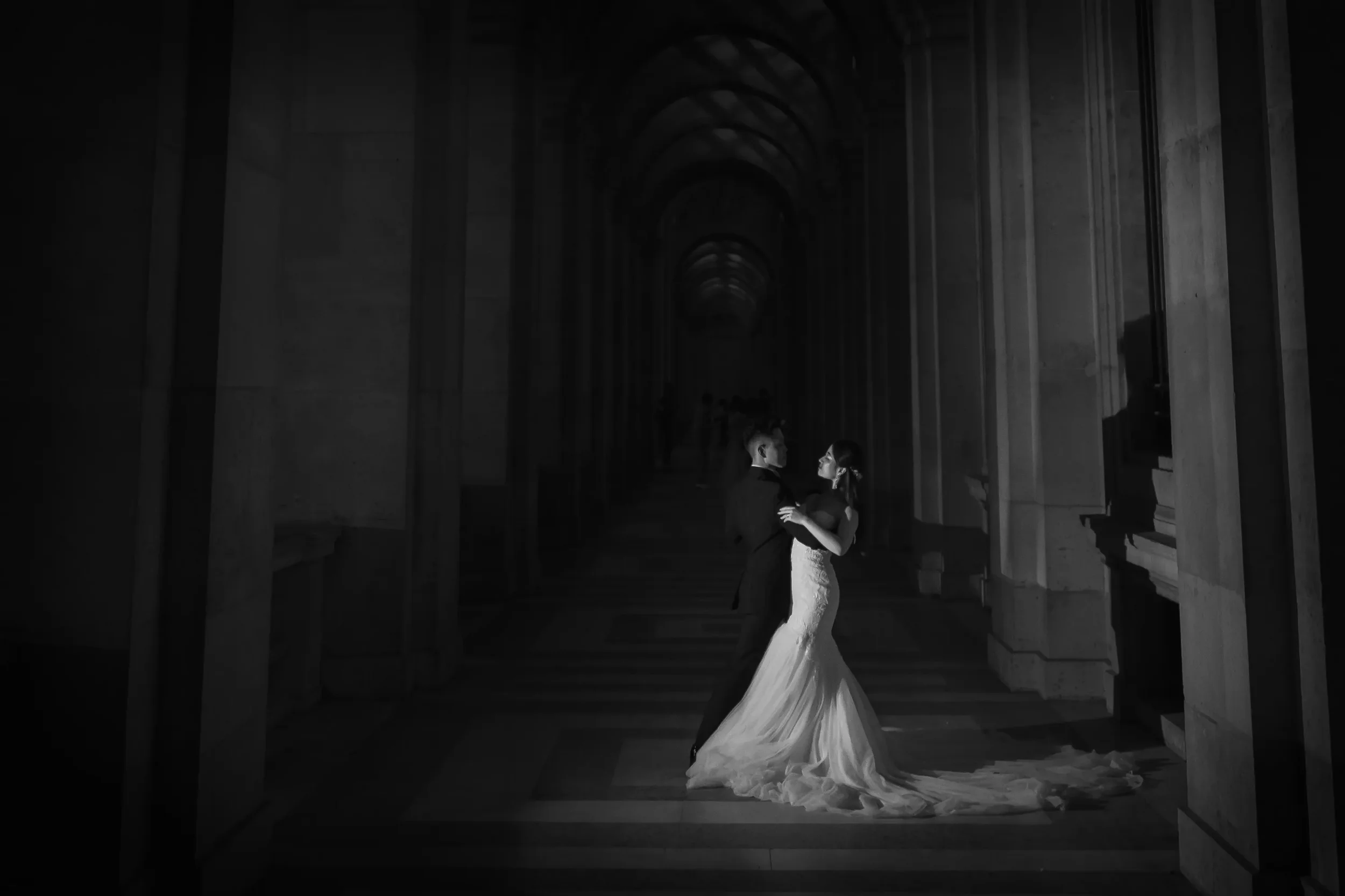 結婚式の衣装を着た新郎新婦がダンスしている黒と白の写真。背景はアーチ型の古い建築物の廊下。