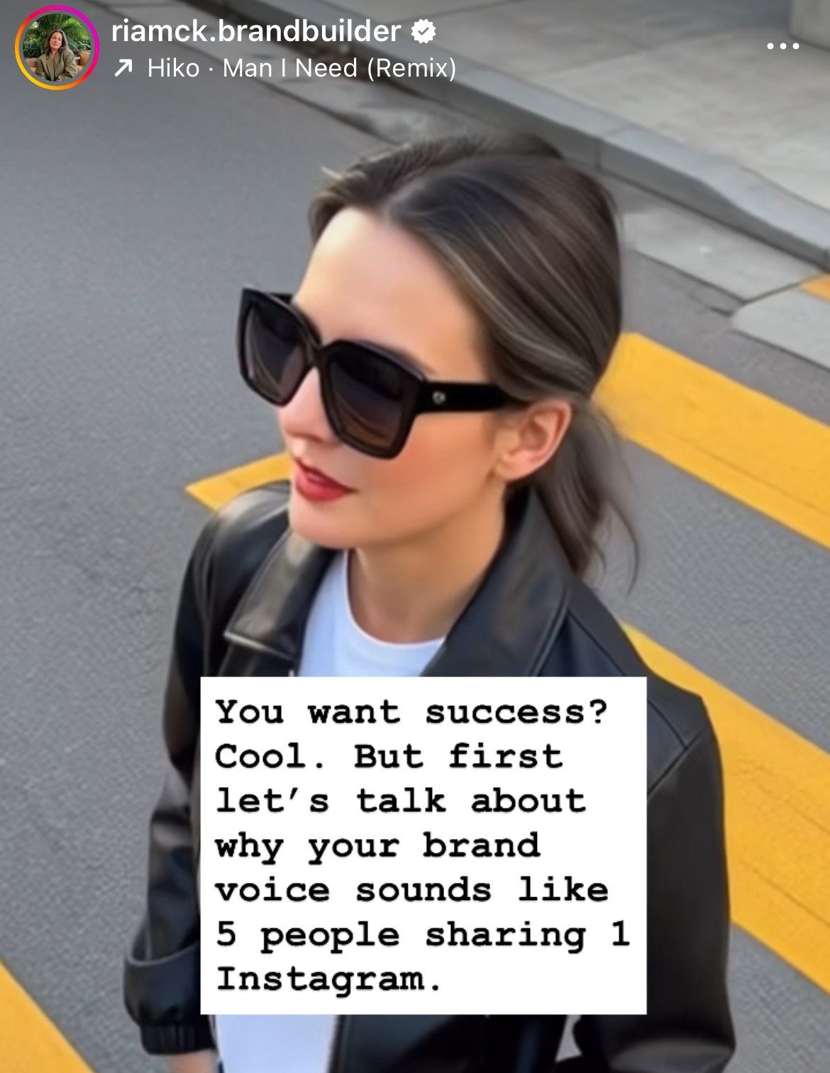 A woman with dark hair, wearing large black sunglasses, a white shirt, and a black leather jacket, standing on a street with yellow road markings.