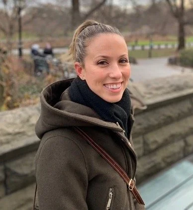 A woman with blonde hair tied in a ponytail, smiling outdoors, wearing a dark coat and a black scarf, standing by a stone wall in a park.