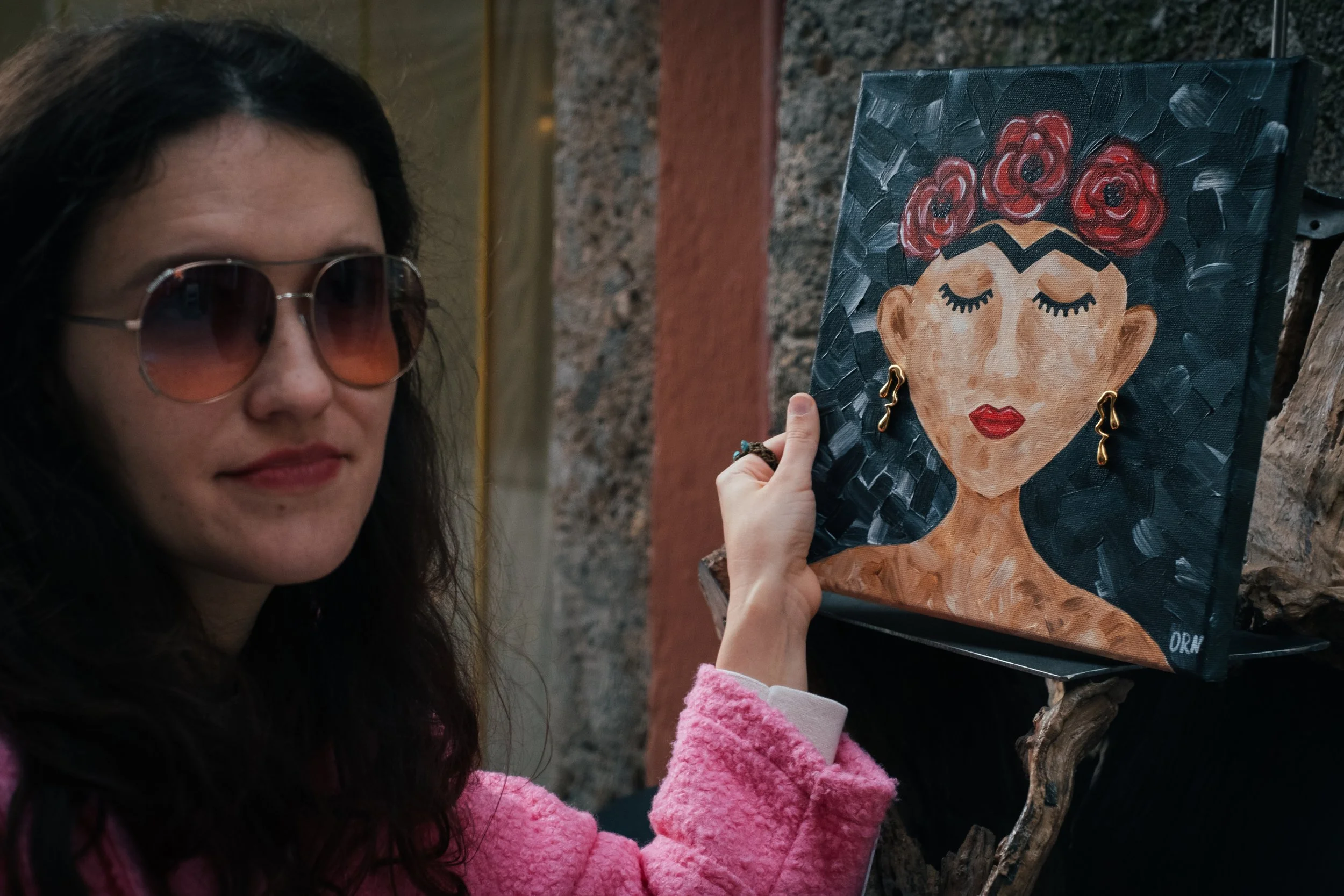 Oleksandra Roche-Newton  in a vibrant pink coat holds a Frida Kahlo portrait painting.