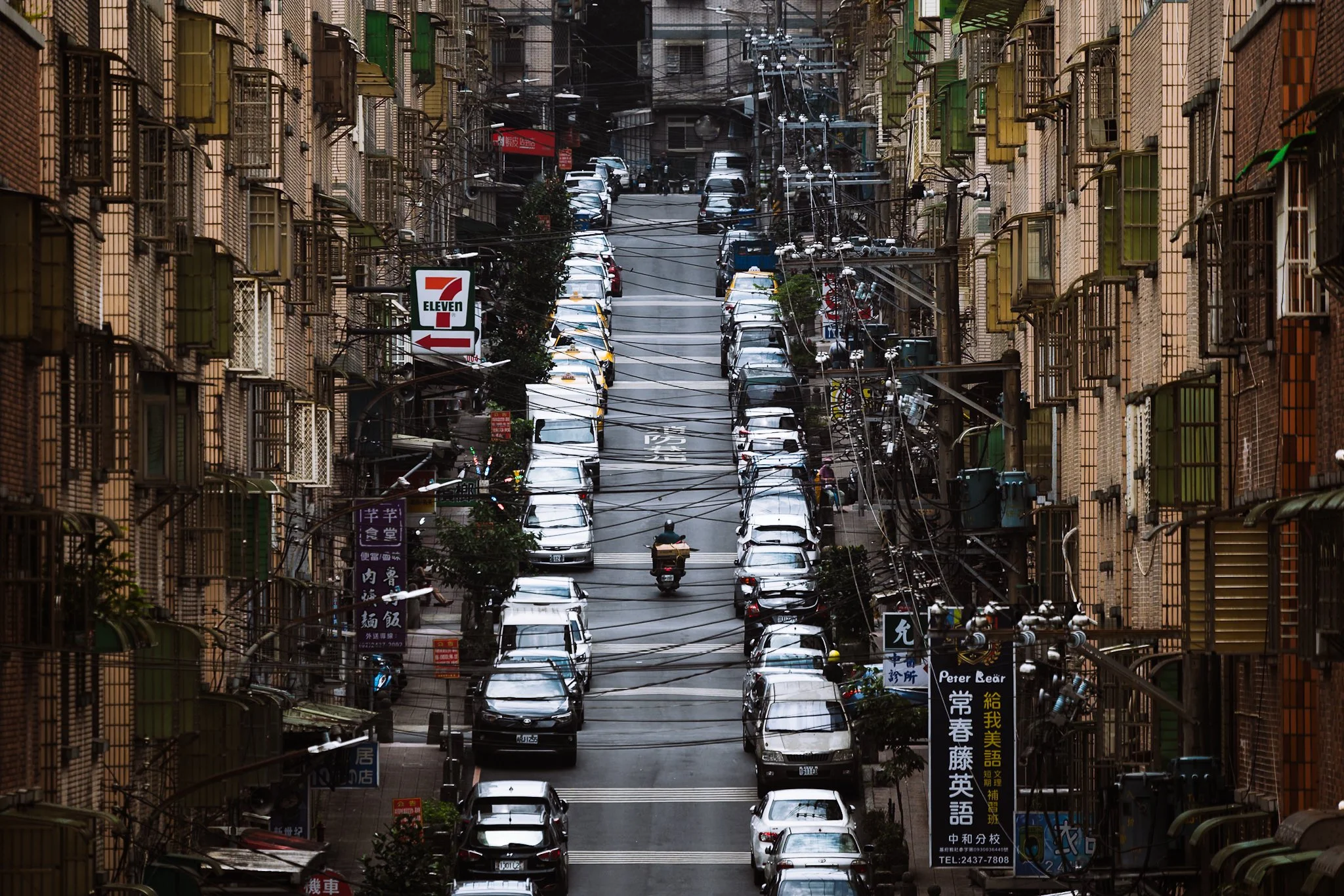 A narrow city street with parked cars on both sides, covered by a web of overhead wires, flanked by multi-story brick buildings with various signs in multiple languages, including English.