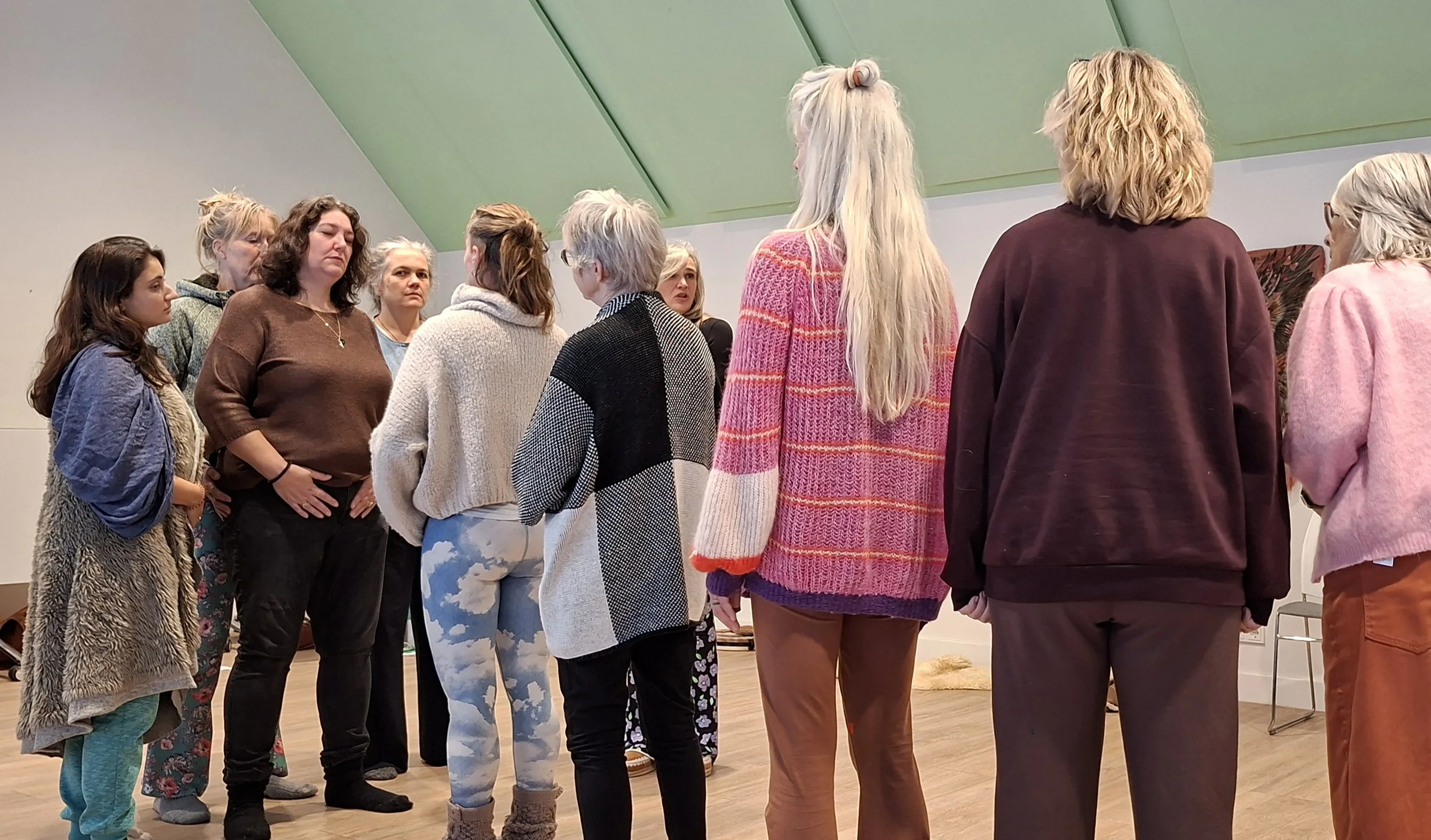 Een groep vrouwen staat in een kring in een kamer met schuine groene plafonds, ze lijken deel te nemen aan een groepsactiviteit of workshop.