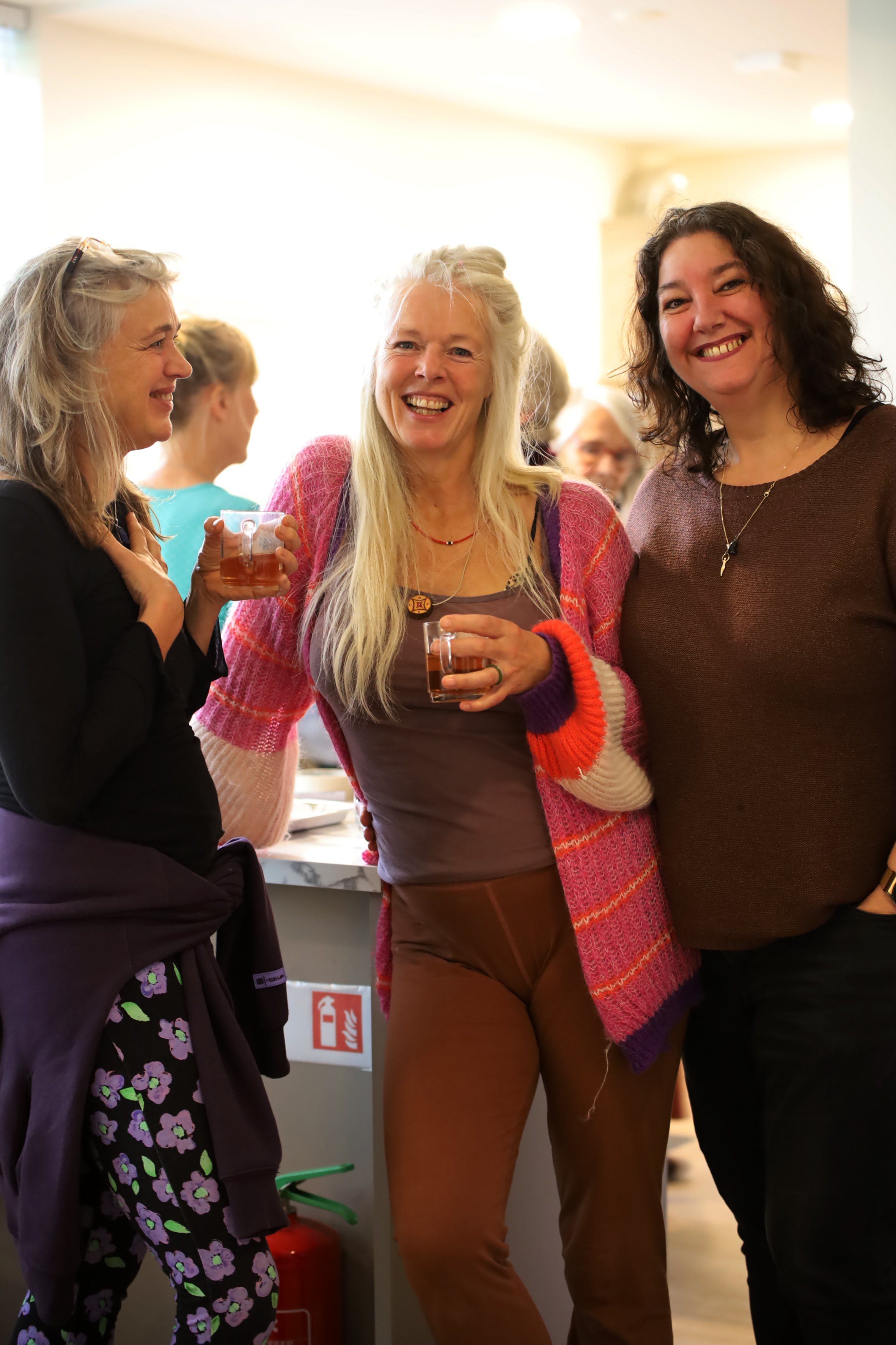 Drie vrouwen lachen en praten samen, met twee glazen drank in hun handen, op een sociale bijeenkomst.
