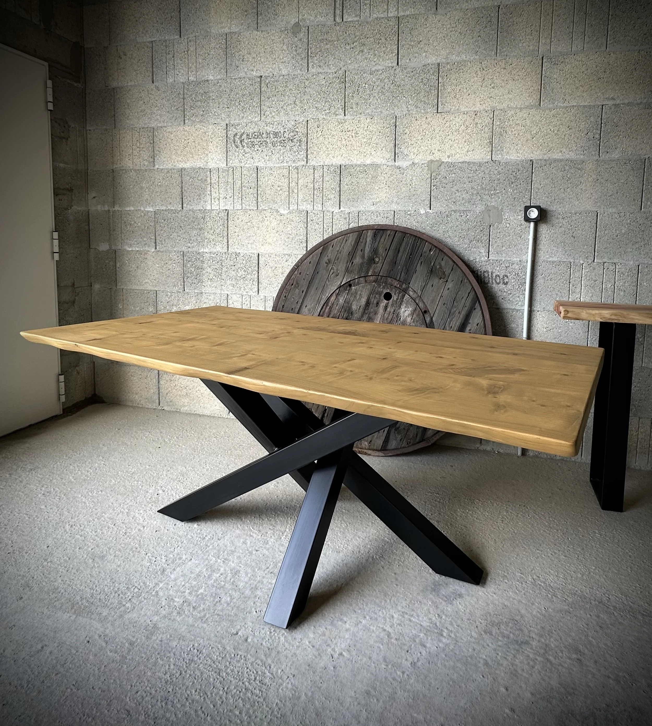 Table en bois avec une base en métal noir, fond en mur en briques gris clair, repose contre une grosse roue en bois rainurée, prise contre le mur.