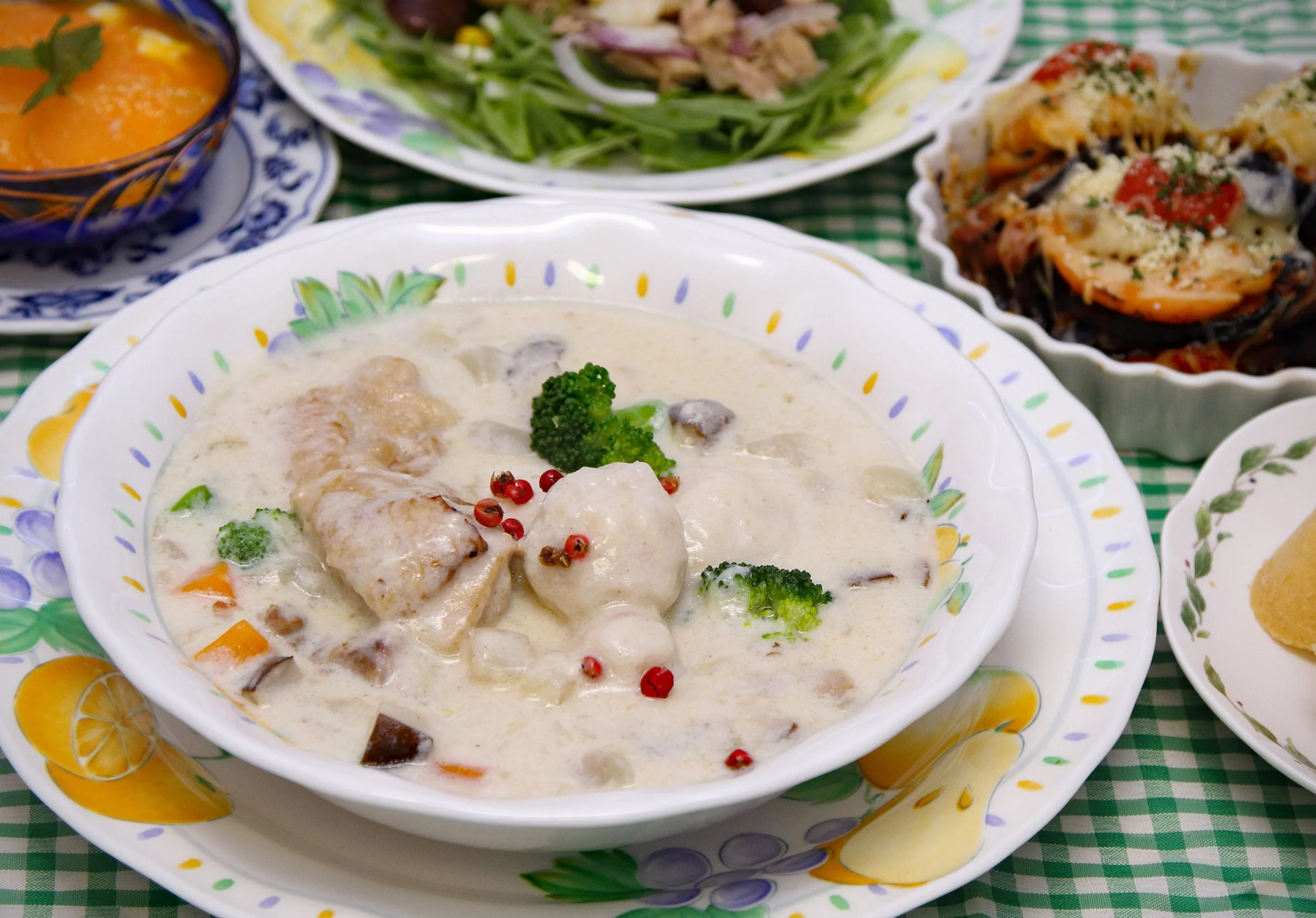 クリーム煮の料理、ブロッコリーとピンクペッパーが飾られている。周囲にサラダ、焼き菓子、蒸しパン、サラダが見える。